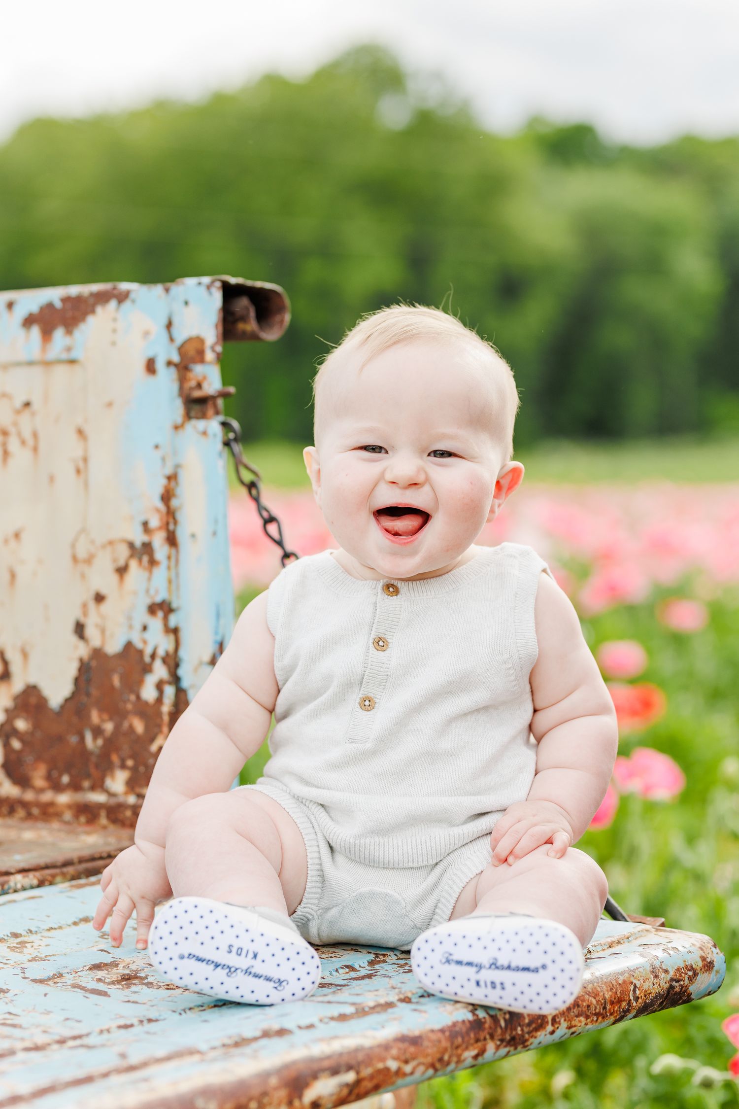 Baby sitting on rustic blue chair in flower field wearing white outfit  in Winston Salem NC  with a Family Photographer