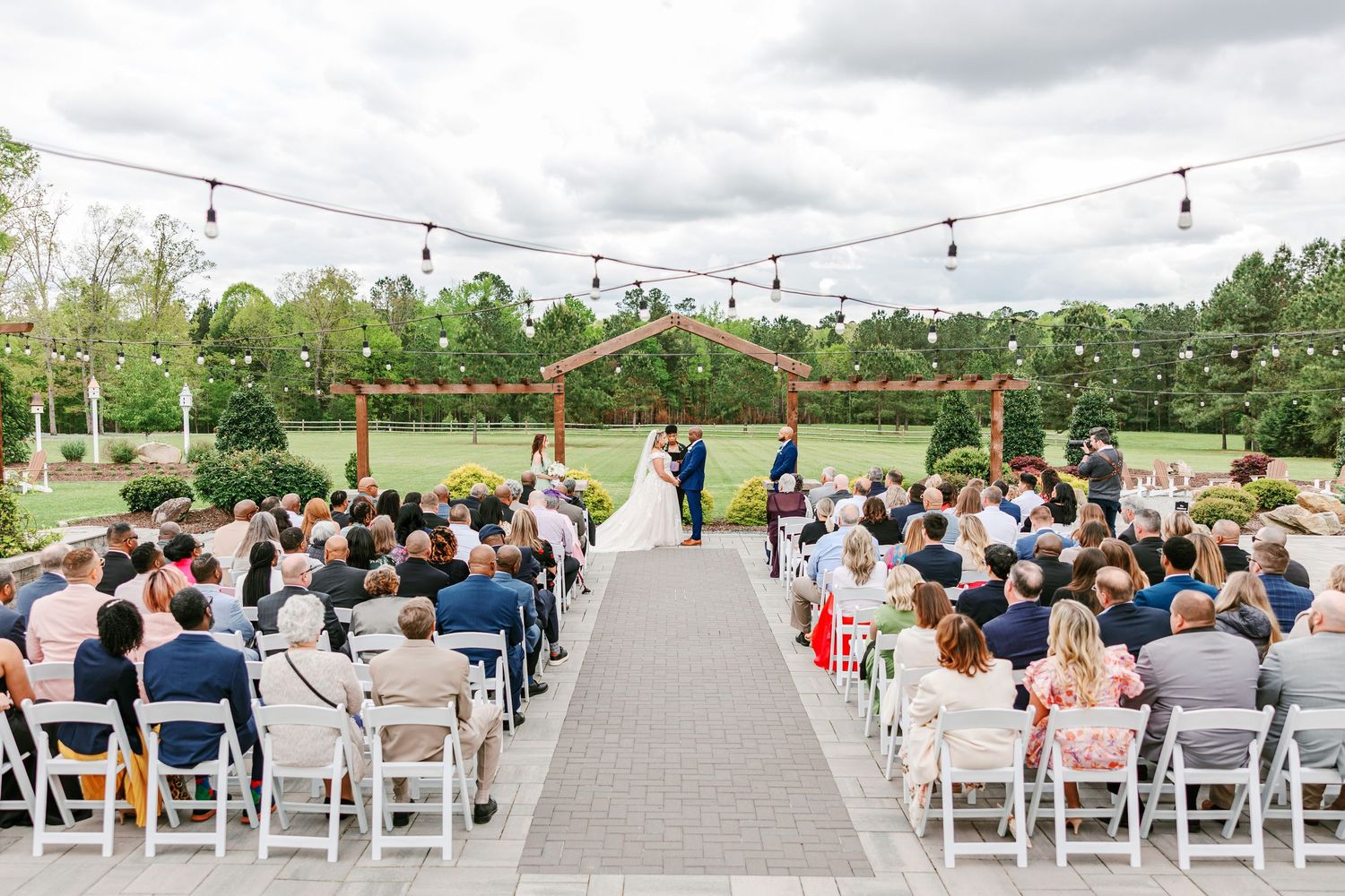 Outdoor wedding ceremony setup with white chairs and string lights under cloudy skies.