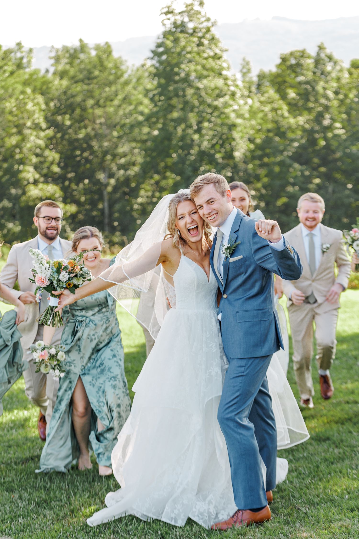 Wedding party celebrates joyfully on a grassy lawn with bridesmaids in blue dresses and groomsmen in light suits at Appalachian View Venue