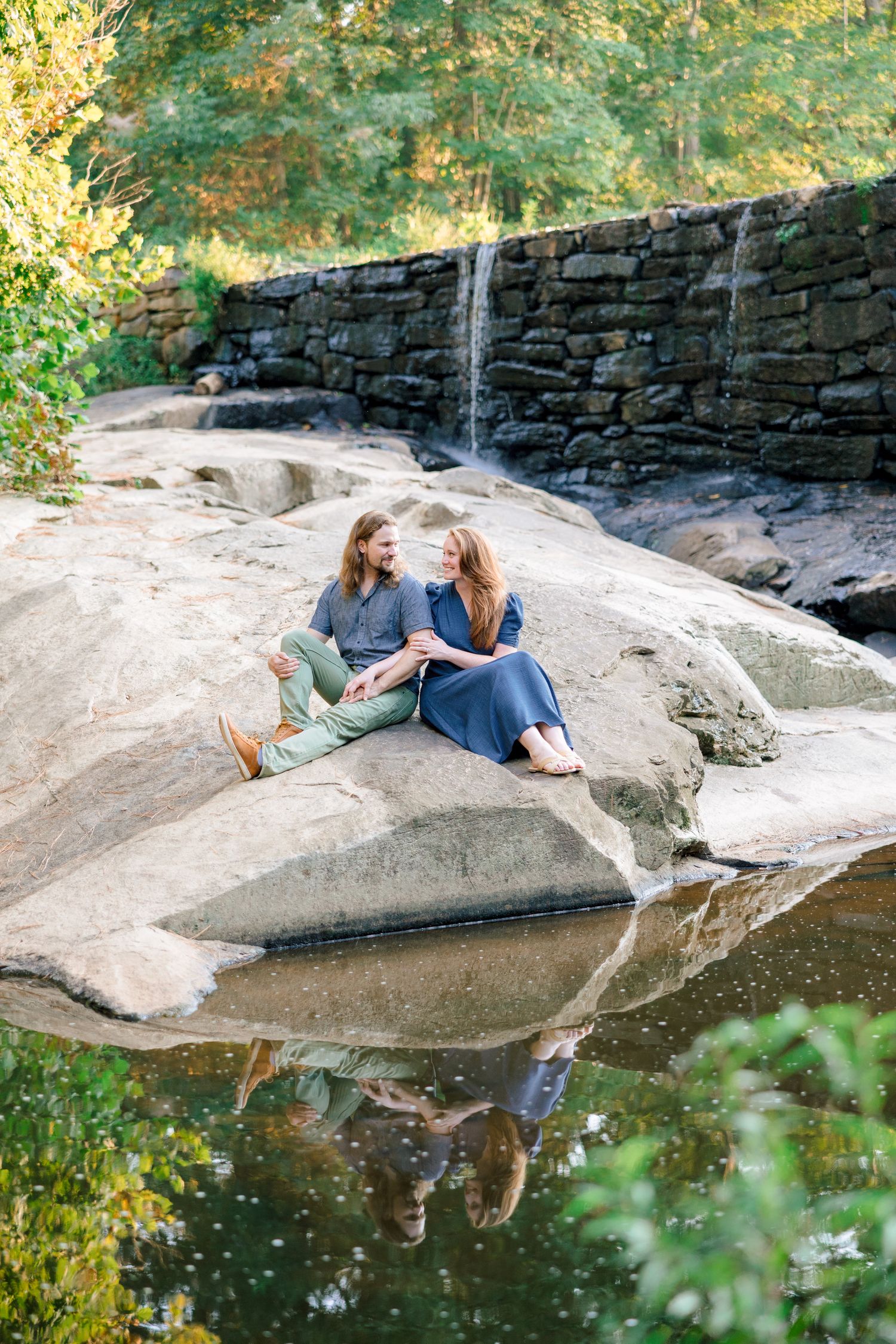 Couple relaxes on large rock formation near stone wall and reflecting pool with Raleigh Engagement Photographer