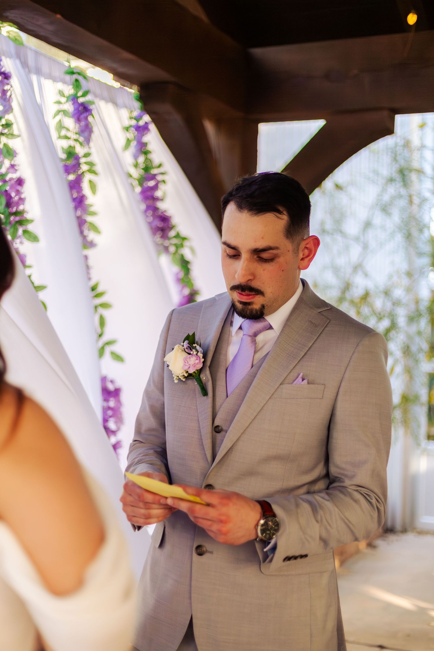 Groom in gray suit reading wedding vows from a card at an outdoor ceremony with purple floral arch backdrop.