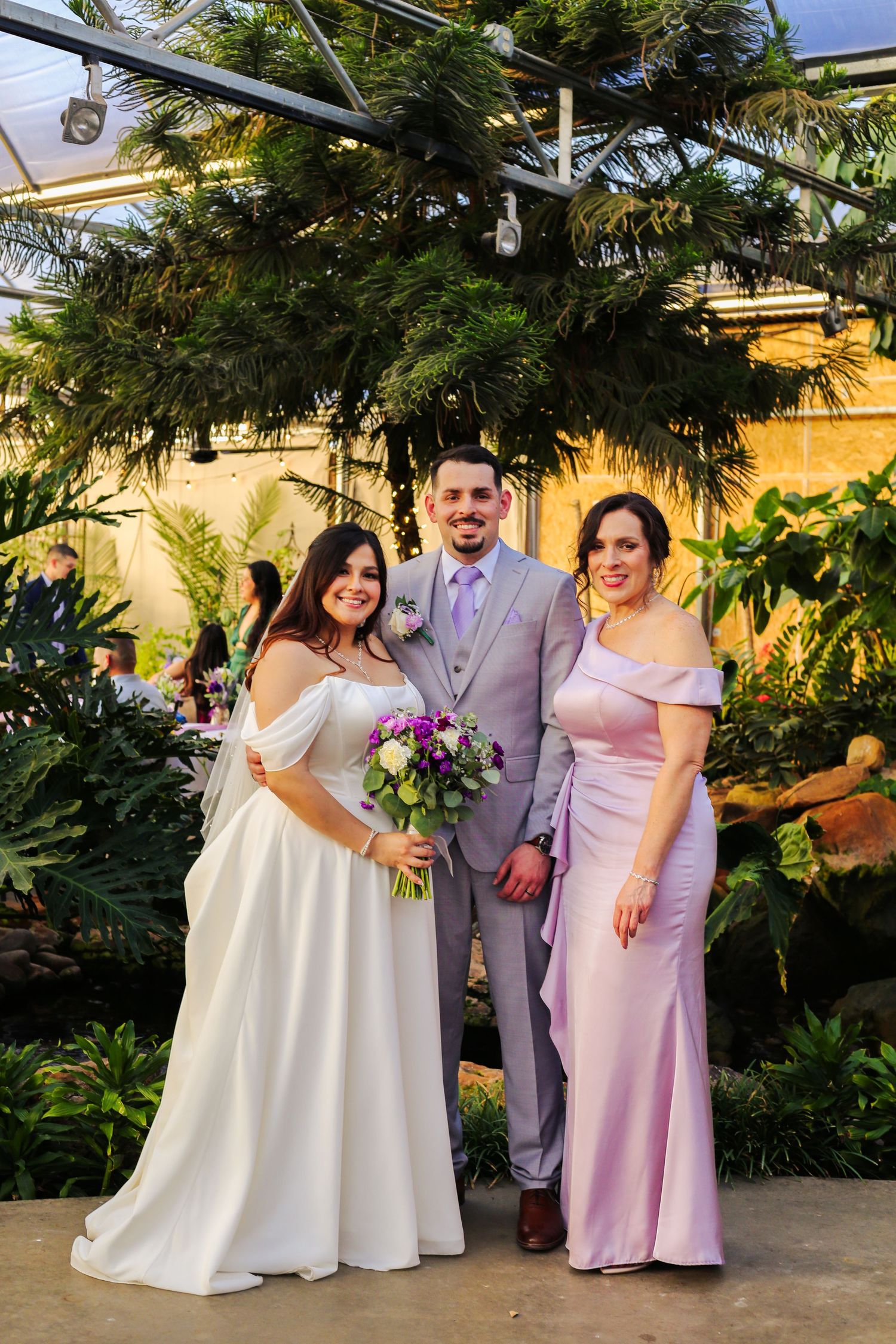 Bride in white gown with groom in gray suit and woman in lavender dress posing in tropical greenhouse.