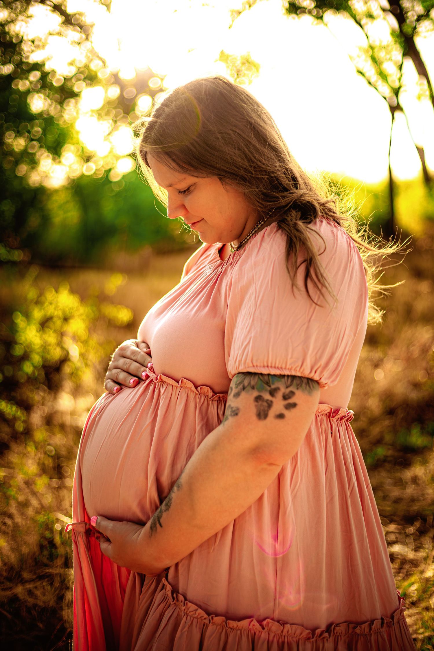 Maternity photos with paw print tattoo in pink dress cradling belly in sunlit field.