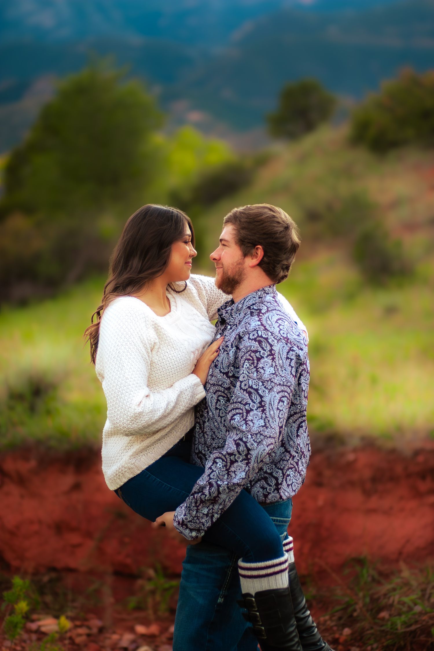 Happy couple embracing and smiling at each other on red dirt trail with lush green hills behind.