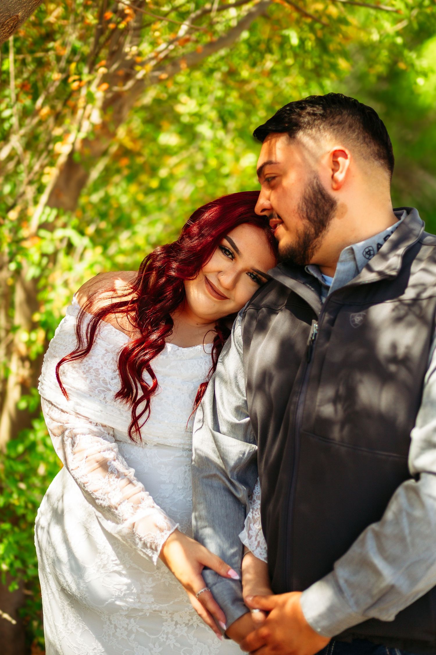 A couple embraces outdoors, woman with red hair in white lace dress leans on bearded man in gray vest, green trees behind.