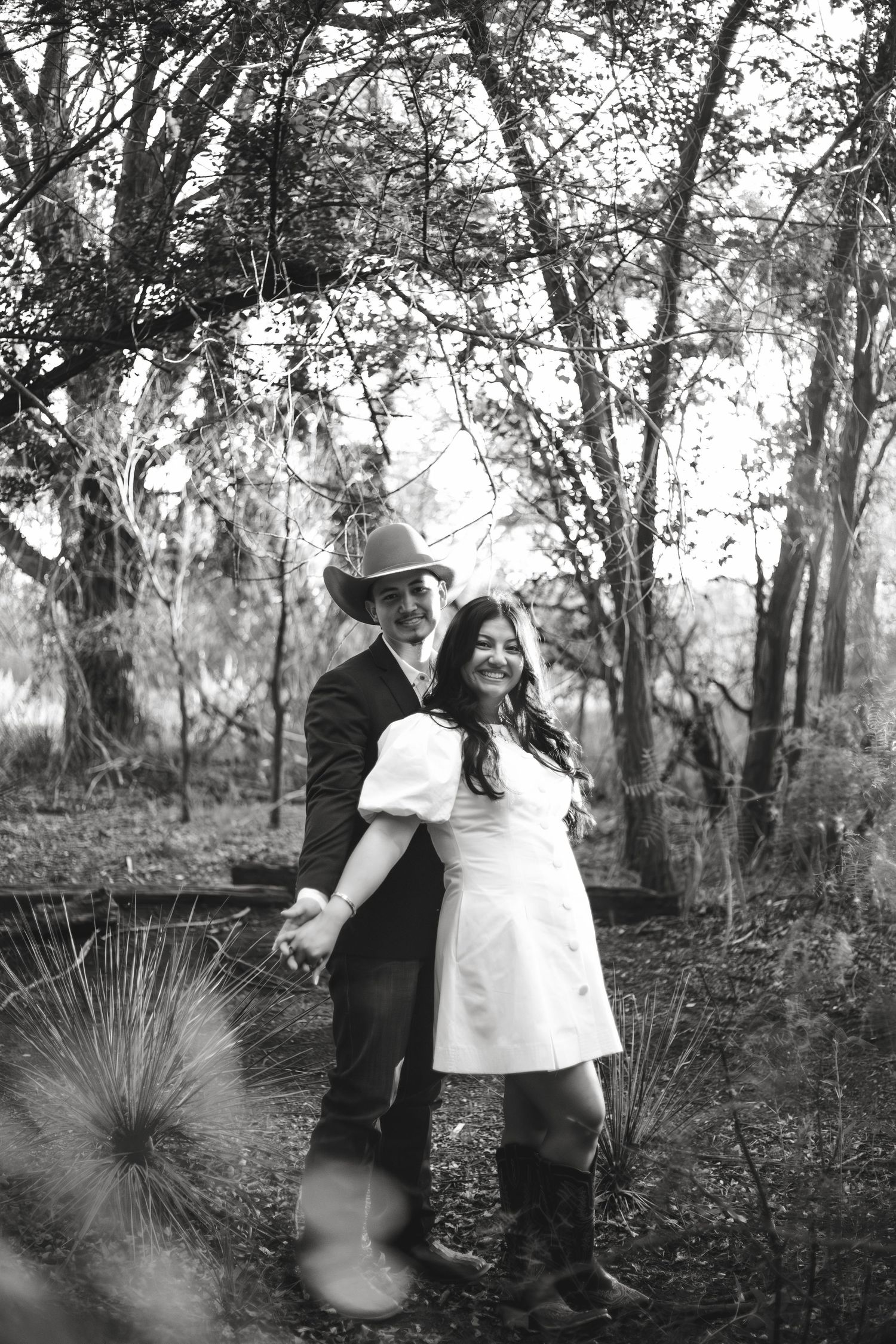 Black and white photo of a smiling couple holding hands in a wooded setting, woman in white dress and man in cowboy hat.