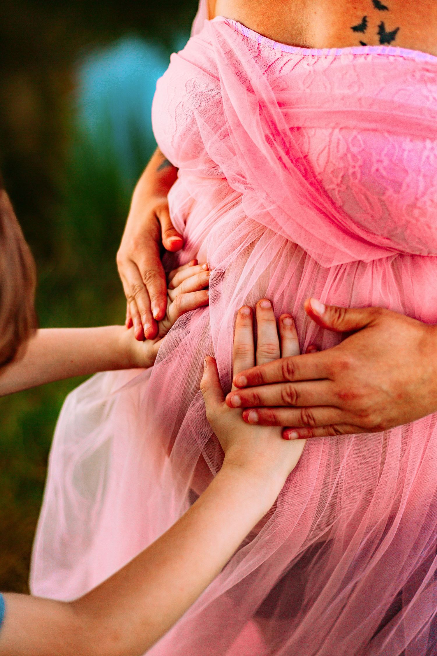 Multiple hands tenderly touching a pregnant woman's belly in a pink tulle maternity dress outdoors.