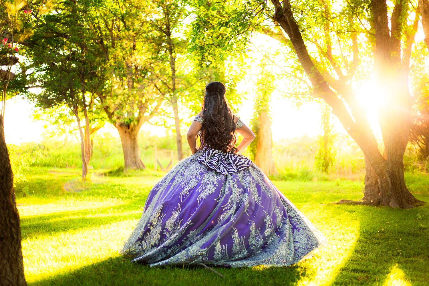 Young woman in purple ball gown standing in sunlit park during golden hour quinceañera celebration.