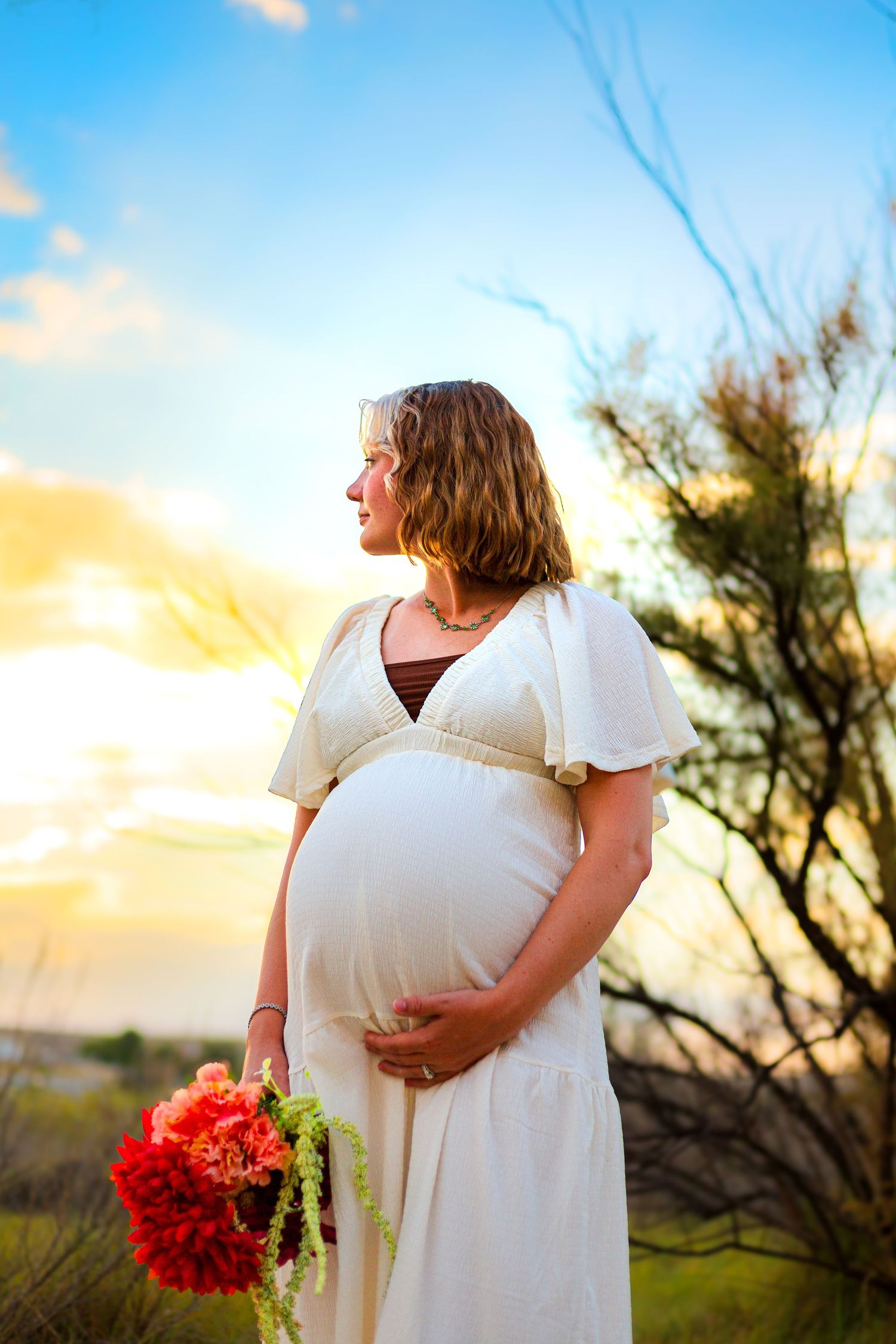 Pregnant woman in white dress holding red flowers at sunset in a natural outdoor setting.