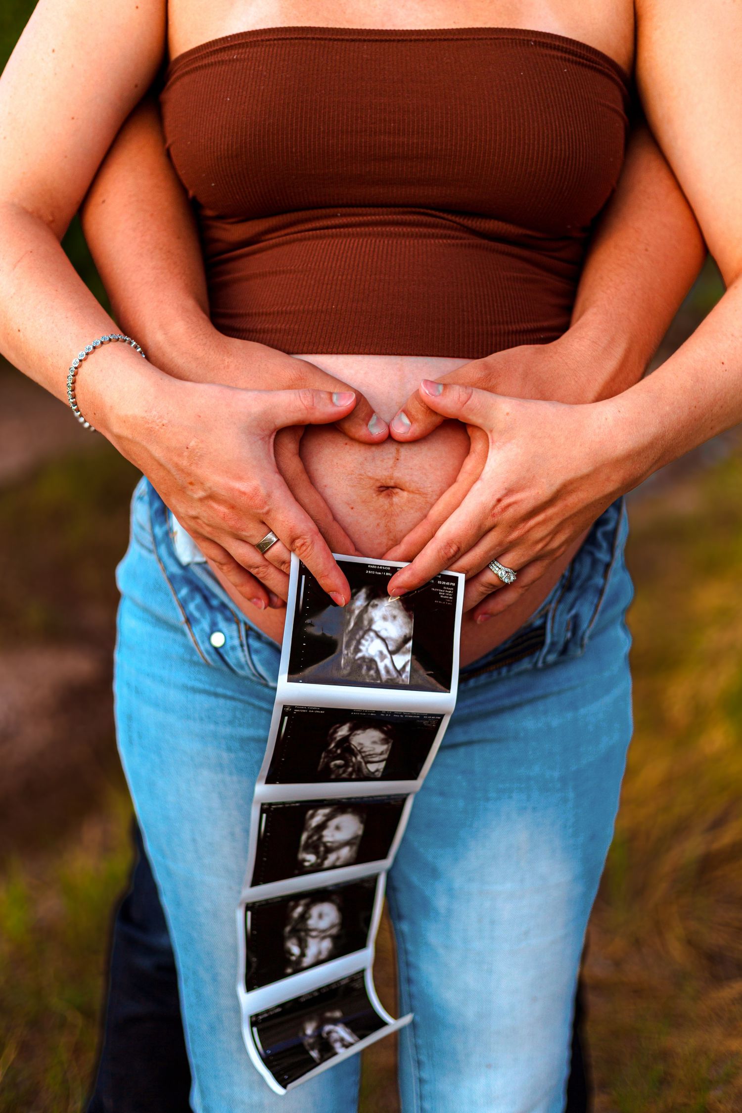 Pregnant woman in brown tube top and jeans forms heart shape on baby bump while holding ultrasound strip photos.