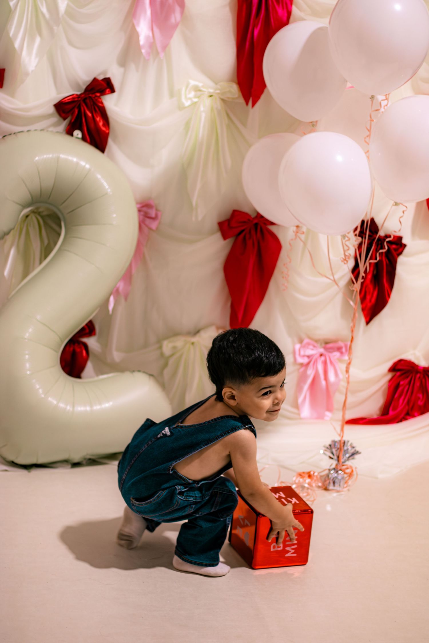 Toddler in denim overalls reaching for gift box at birthday party.