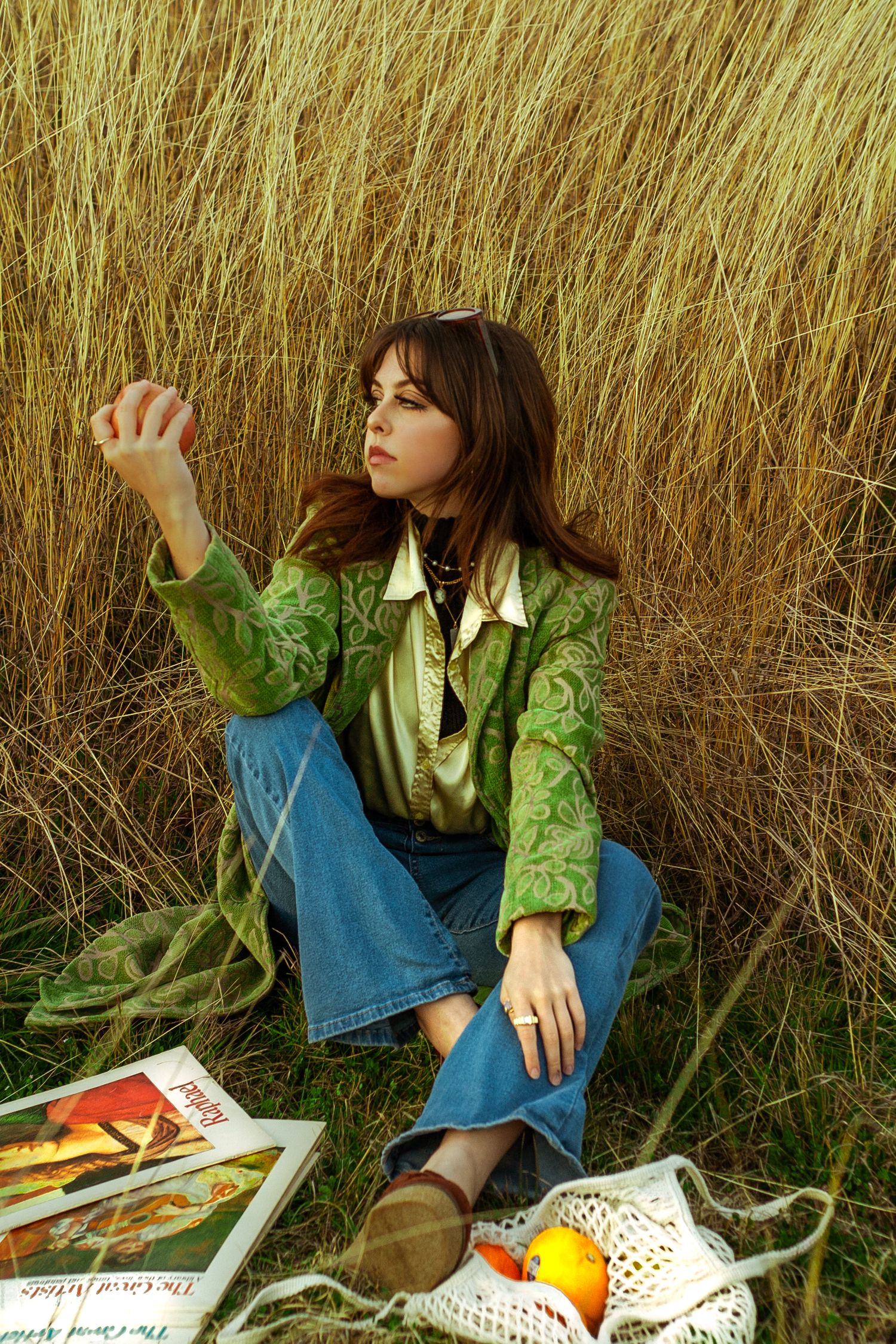 Brunette woman in green jacket sitting in tall grass with books and fruit.