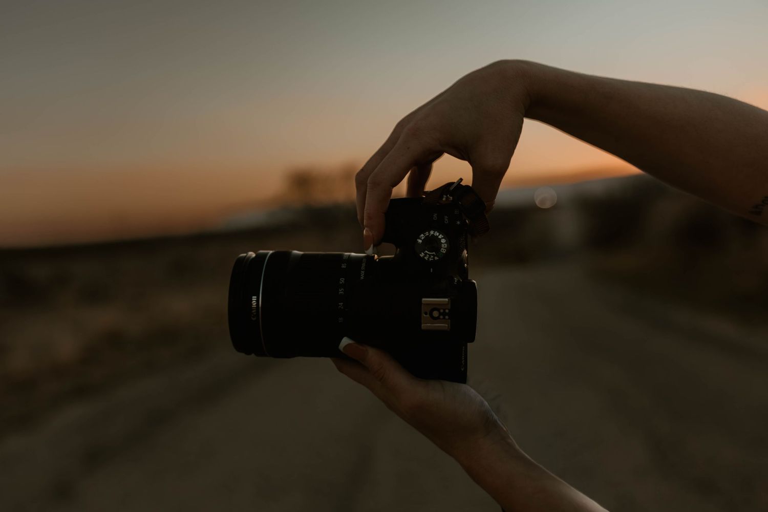 Hands holding a black DSLR camera against a dramatic dusk sky on an empty road.