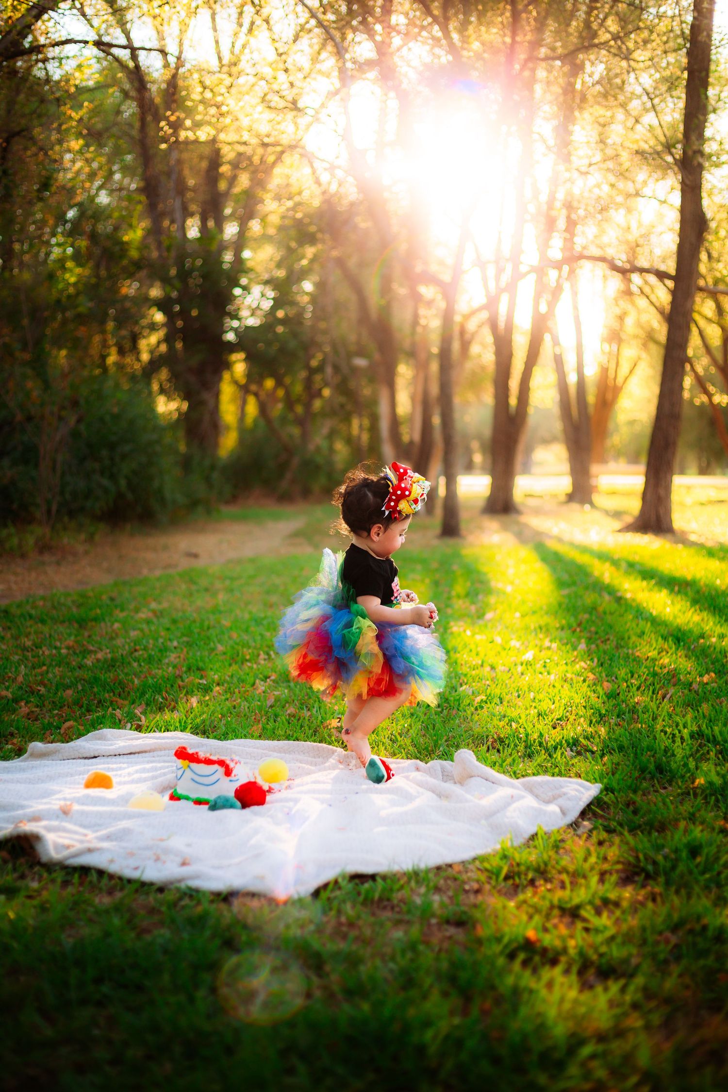 Toddler girl in rainbow tutu dancing on white blanket in sunlit park during golden hour.