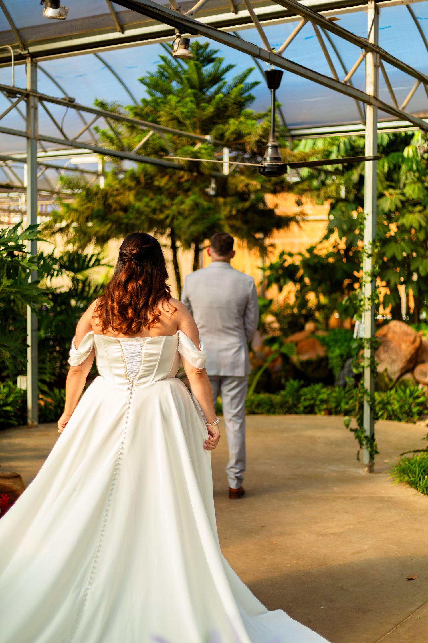 Bride in white off-shoulder gown and groom in grey suit walk hand-in-hand through a lush greenhouse wedding venue.