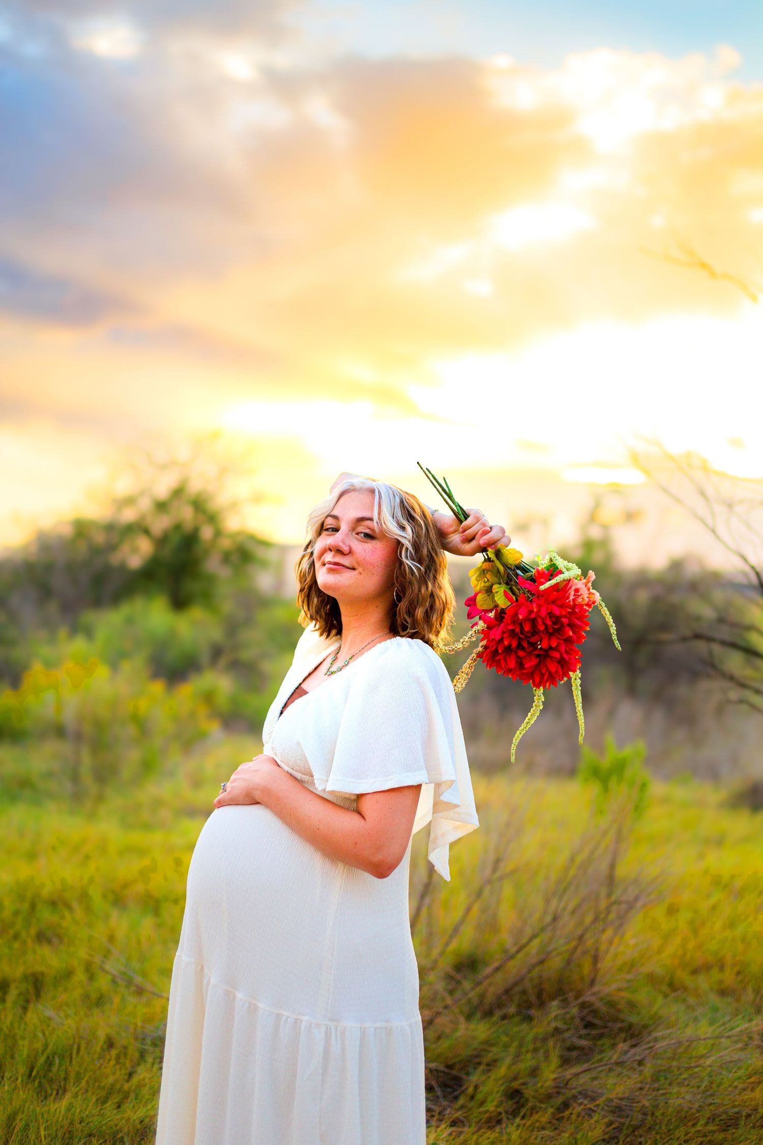 Pregnant woman in white dress holding red flowers at sunset in a field, wearing a floral crown.
