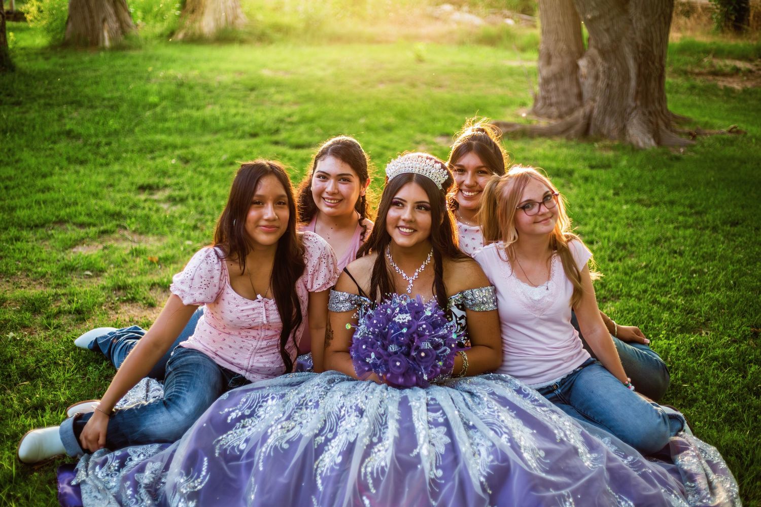 Five young women pose outdoors on green grass, one wearing a purple quinceañera gown and tiara holding a floral bouquet.