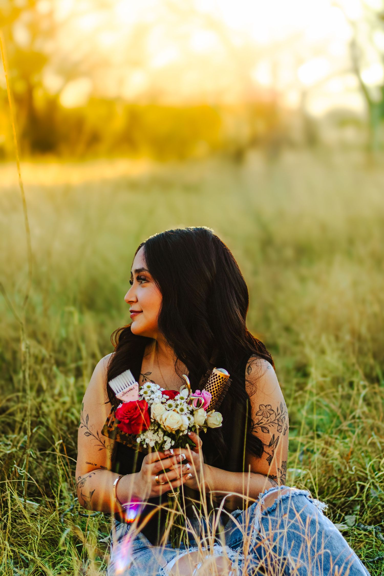 Woman with tattoos sits in golden field at sunset, holding colorful wildflower bouquet, smiling peacefully.