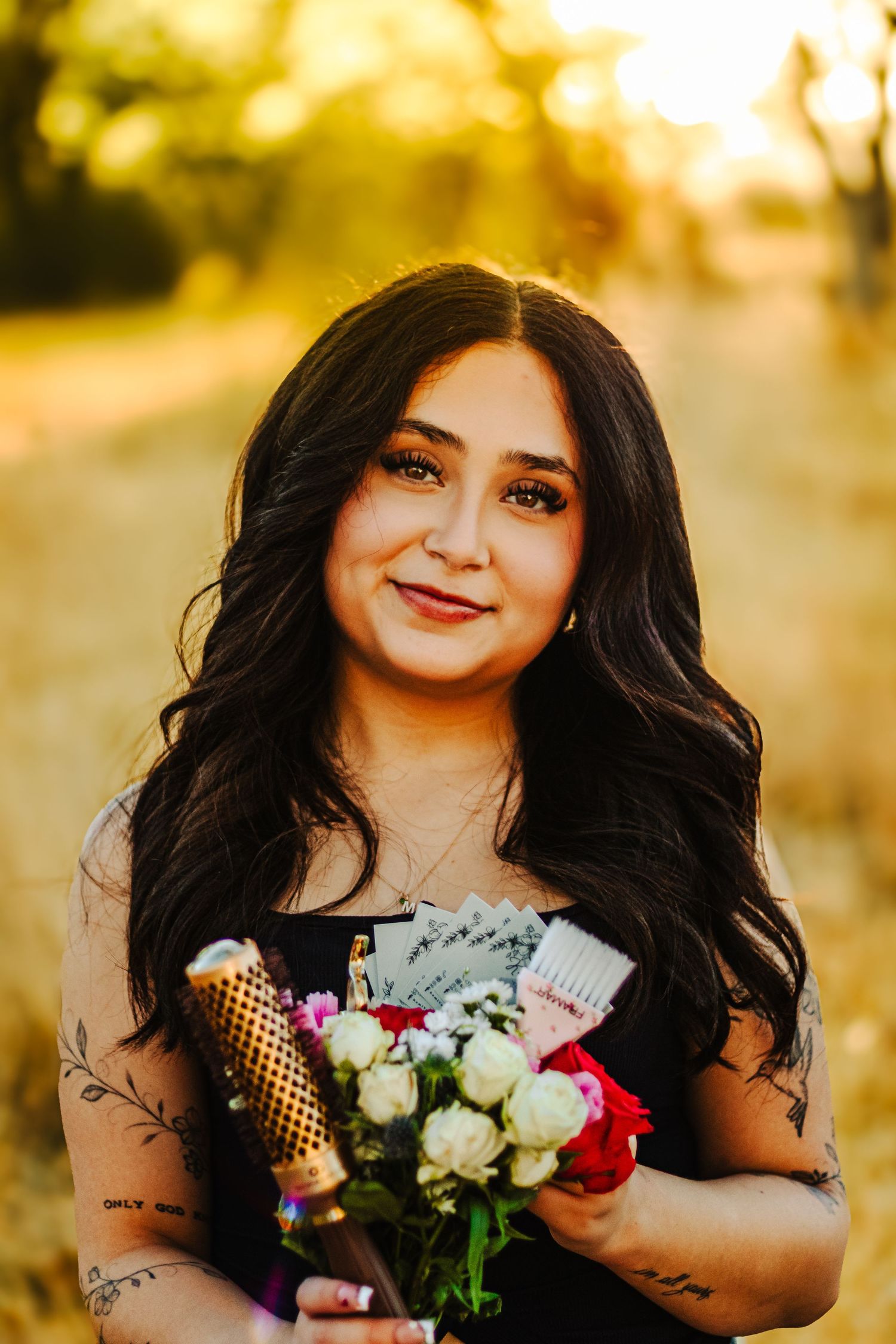 Young woman with long dark hair holds flowers and champagne bottle outdoors in golden sunset light.