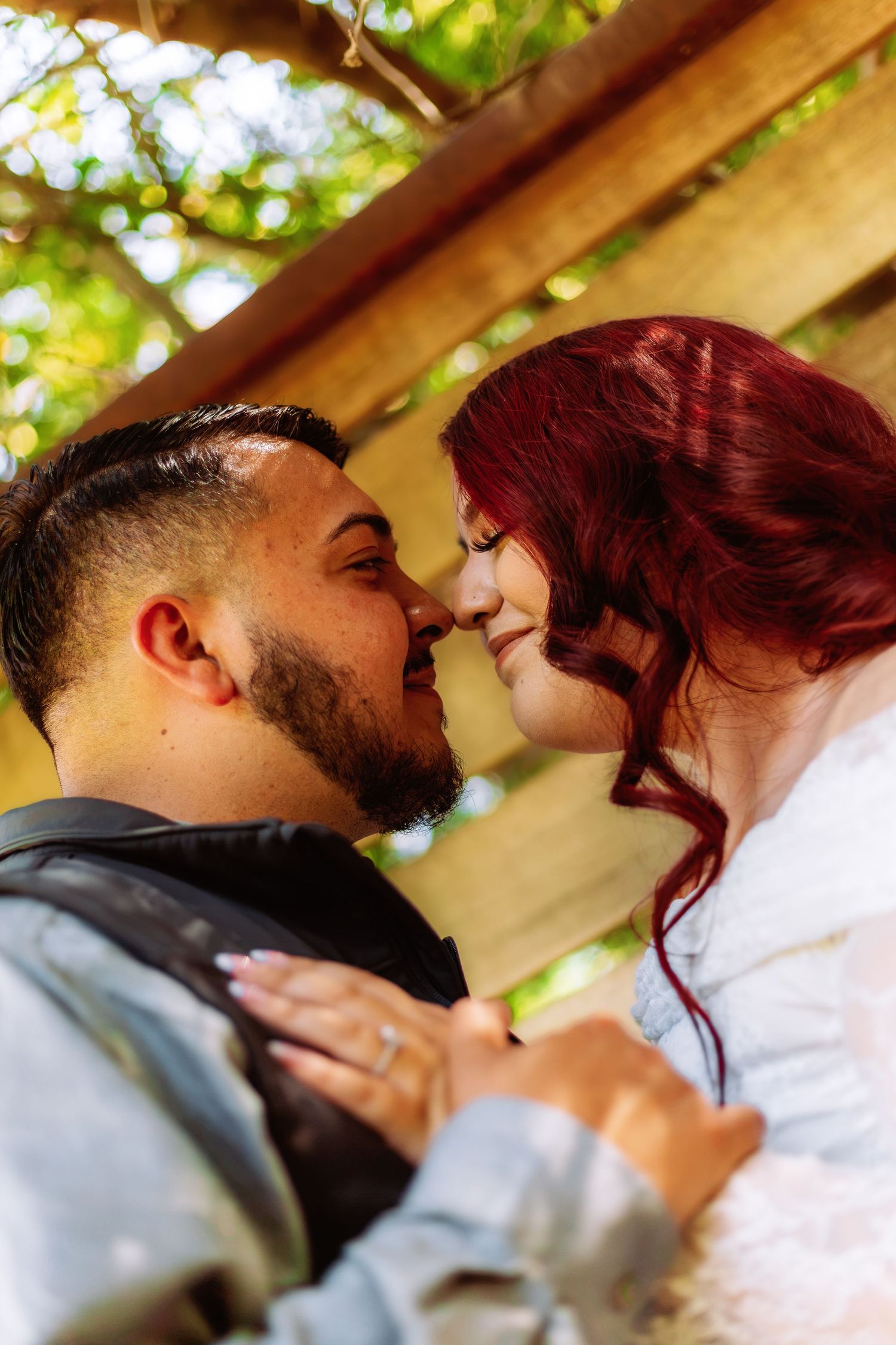 A couple shares a romantic moment, nearly kissing under a wooden pergola surrounded by lush greenery.