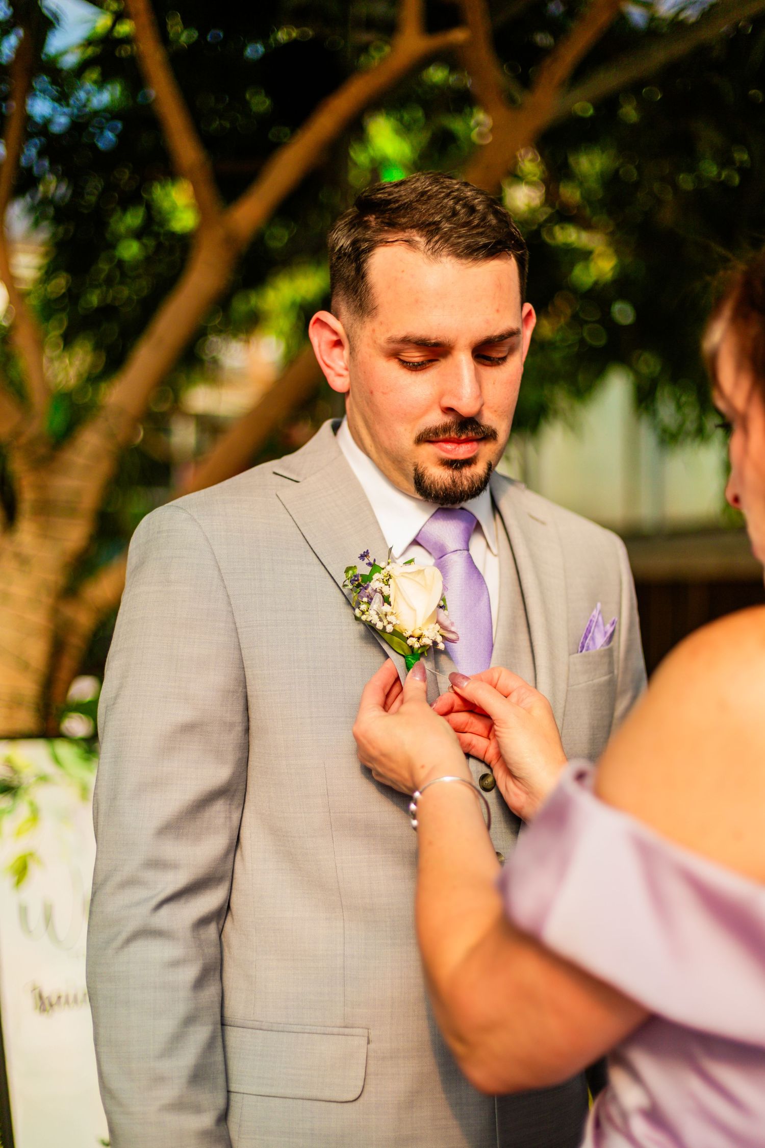 A groom in a gray suit with lavender tie has a white boutonniere pinned to his lapel by a woman in purple.