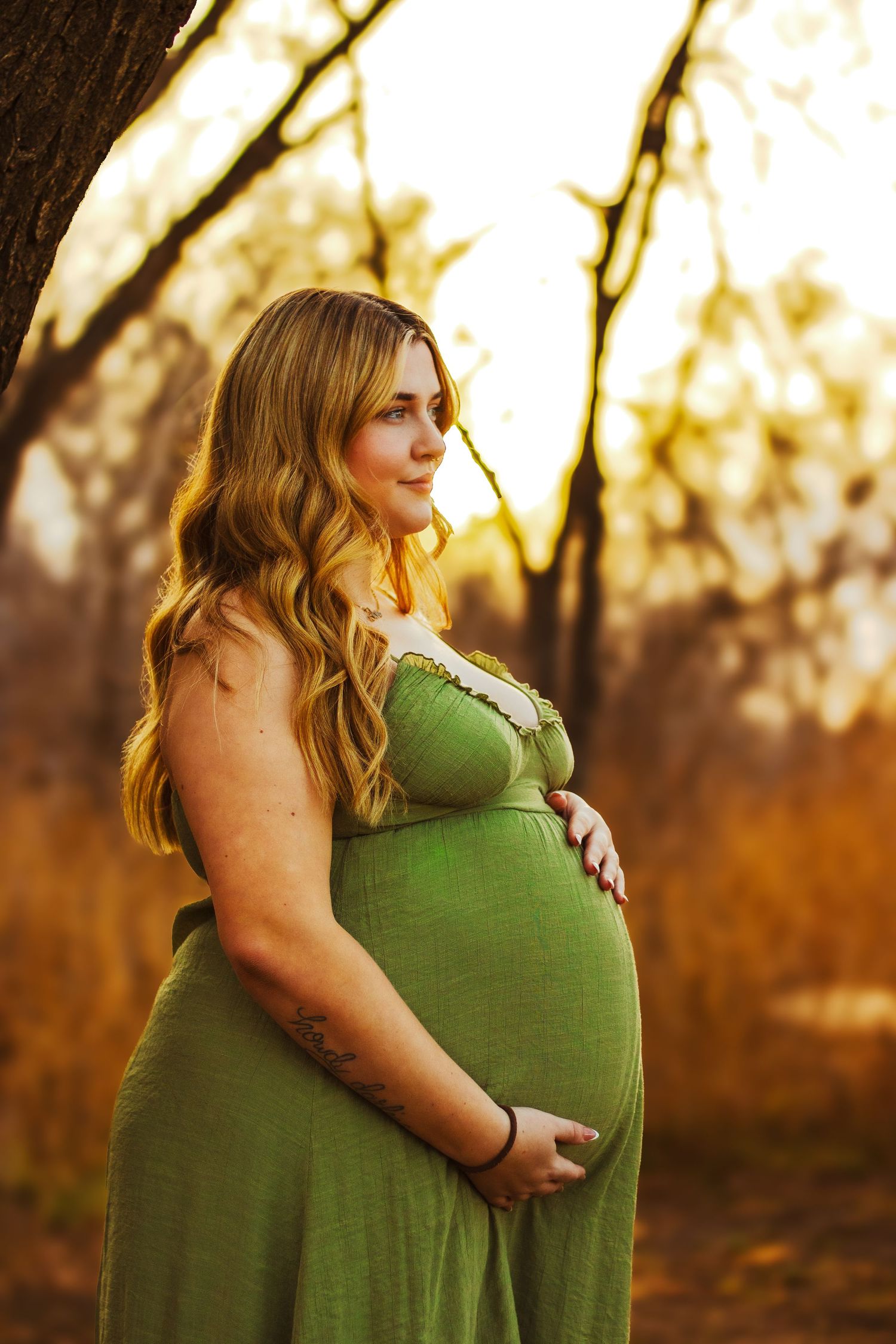 Pregnant woman in green dress cradling baby bump in golden sunset light.