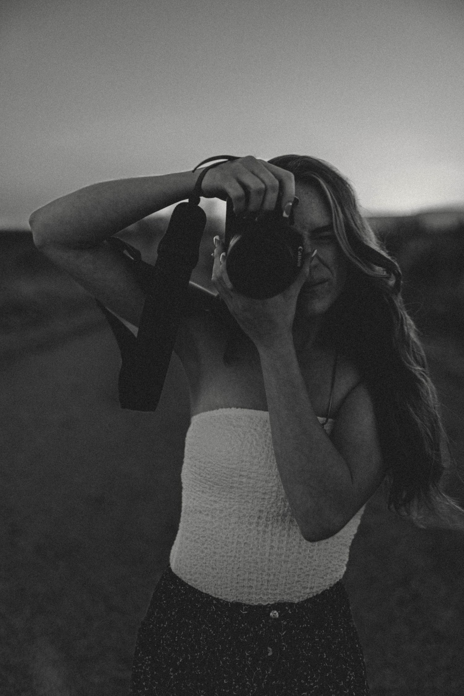 Black and white photo of a woman holding a camera up to shoot at twilight on an open road.