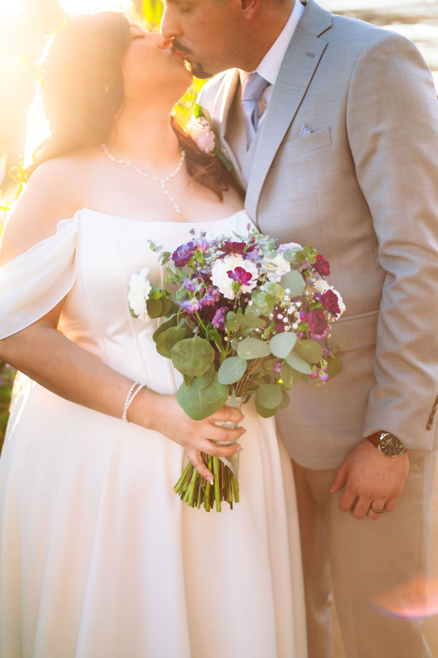 Newlyweds sharing a romantic kiss in golden sunlight, bride holding a purple and white floral bouquet.