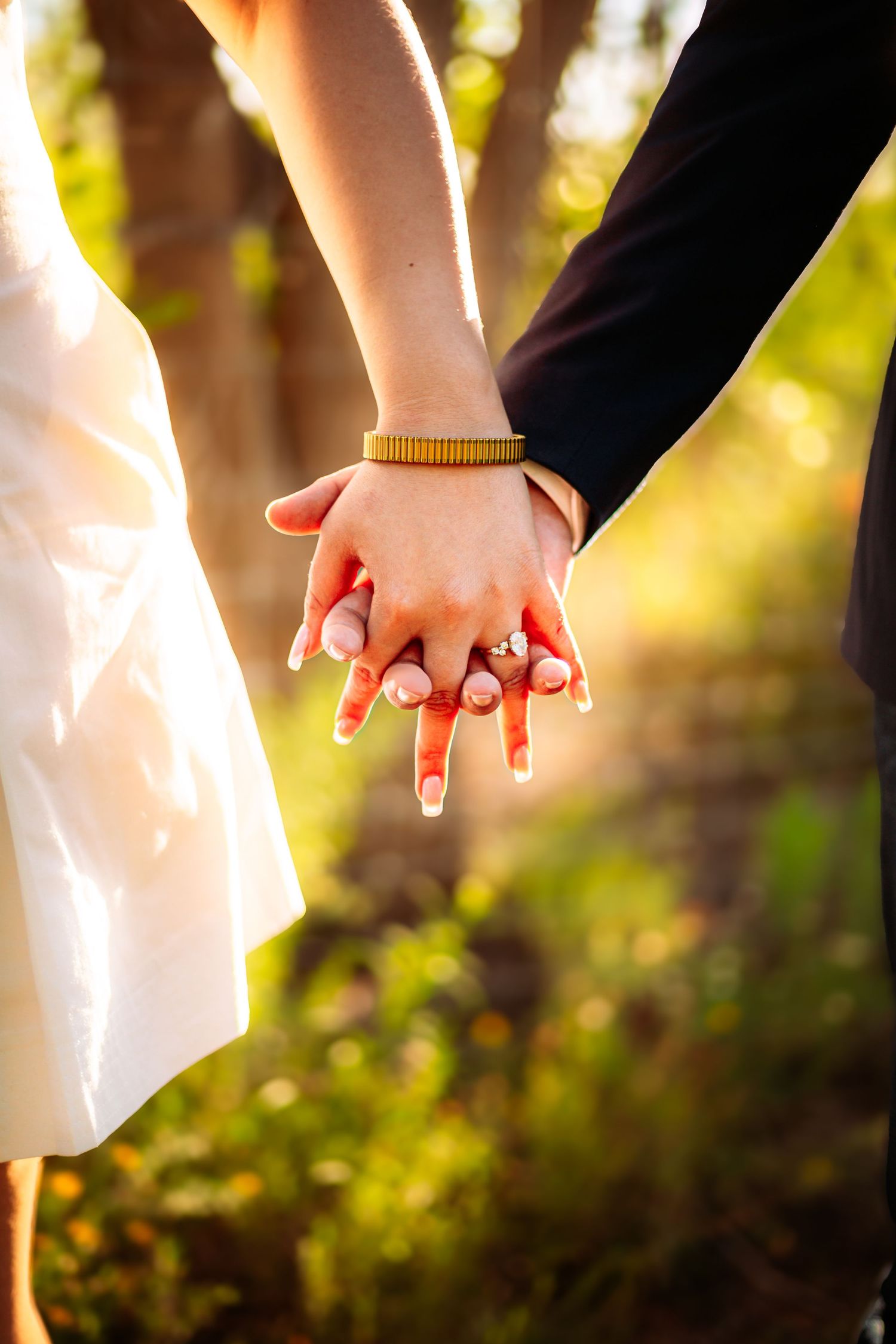 Close-up of a couple holding hands, showing an engagement ring and gold bracelet, with a sunlit forest background.