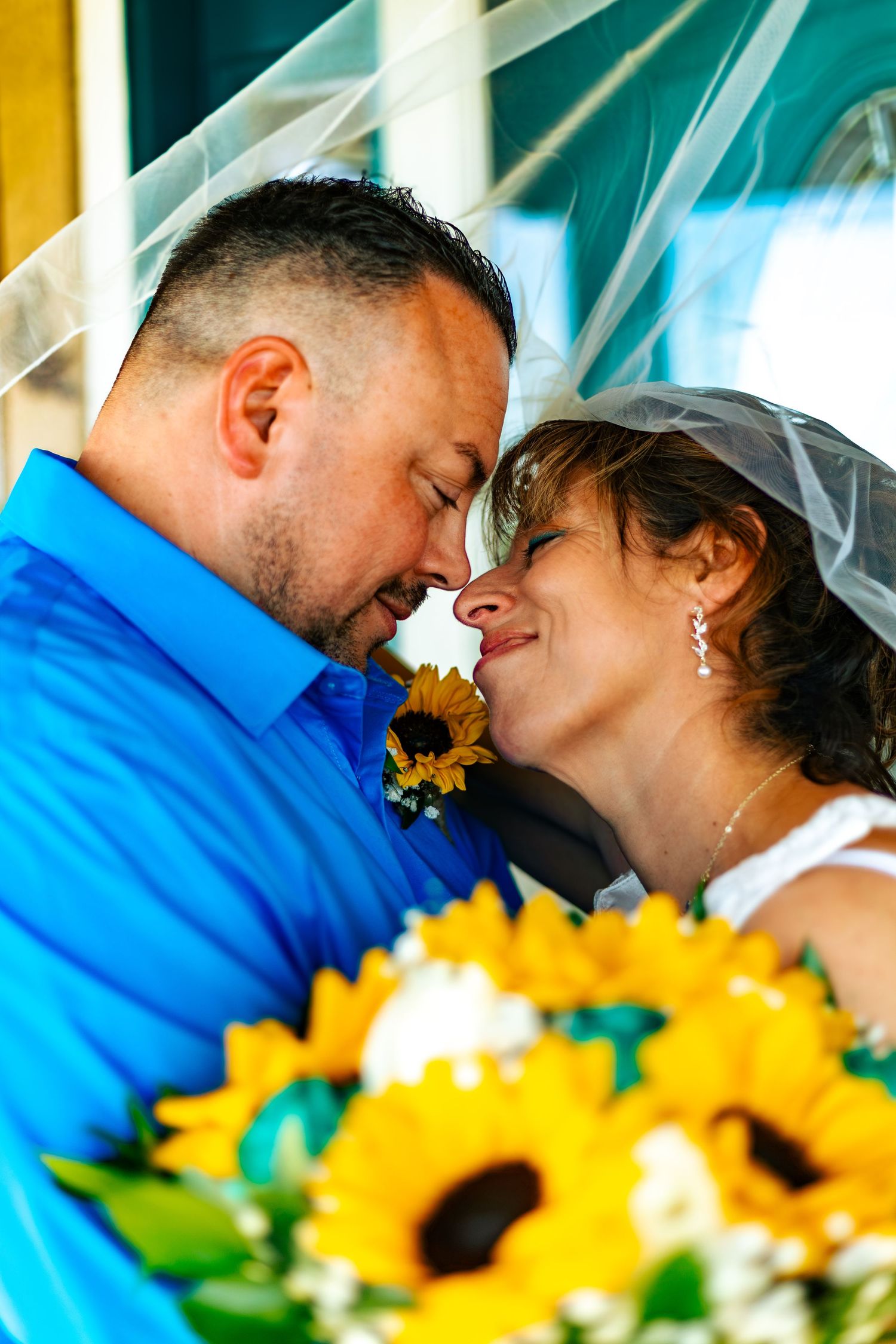 Bride and groom share intimate forehead touch under veil with sunflower bouquet in foreground.