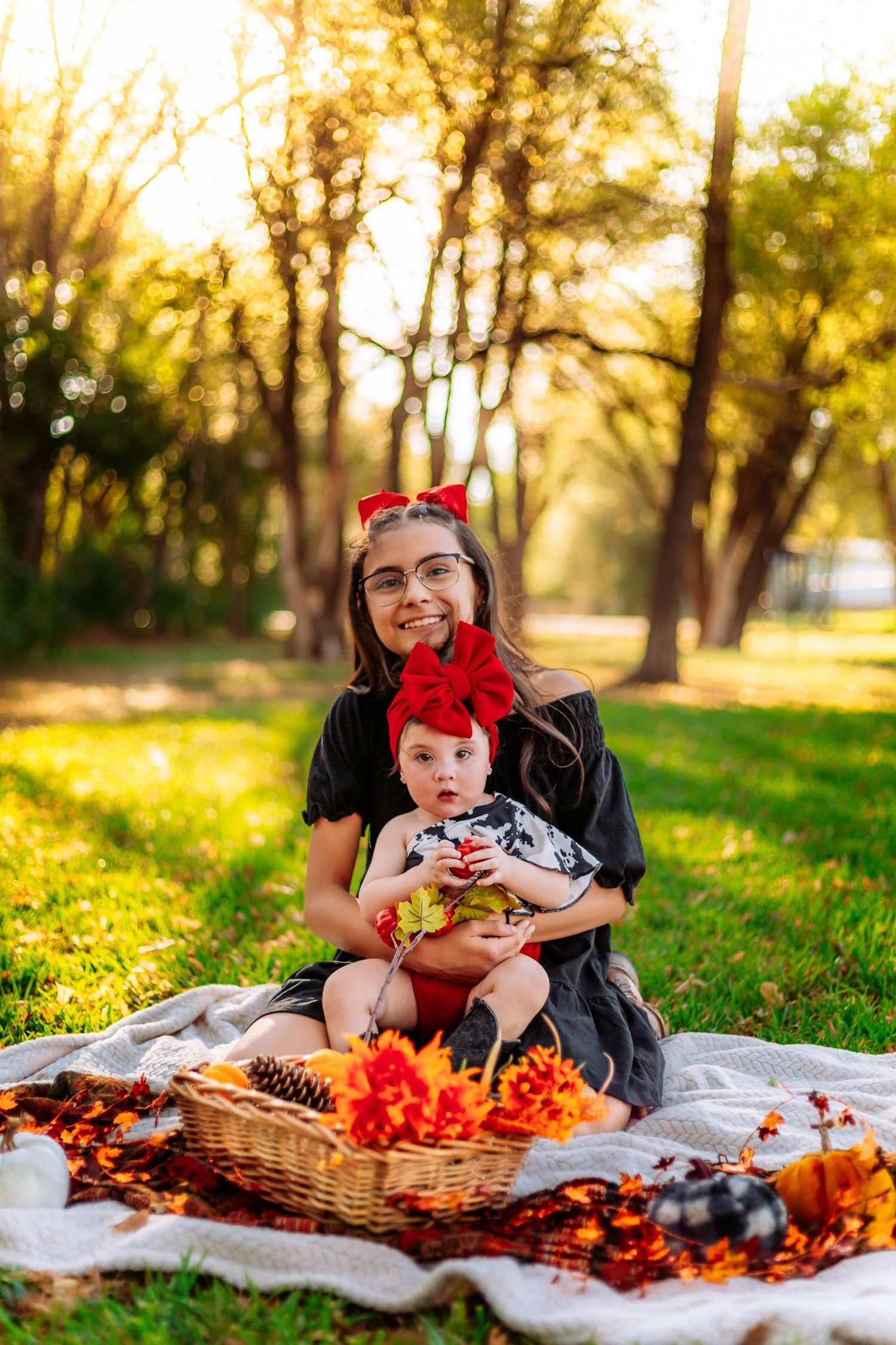 A young woman and baby in matching red bows sit on a blanket surrounded by fall decor in a sunny park.