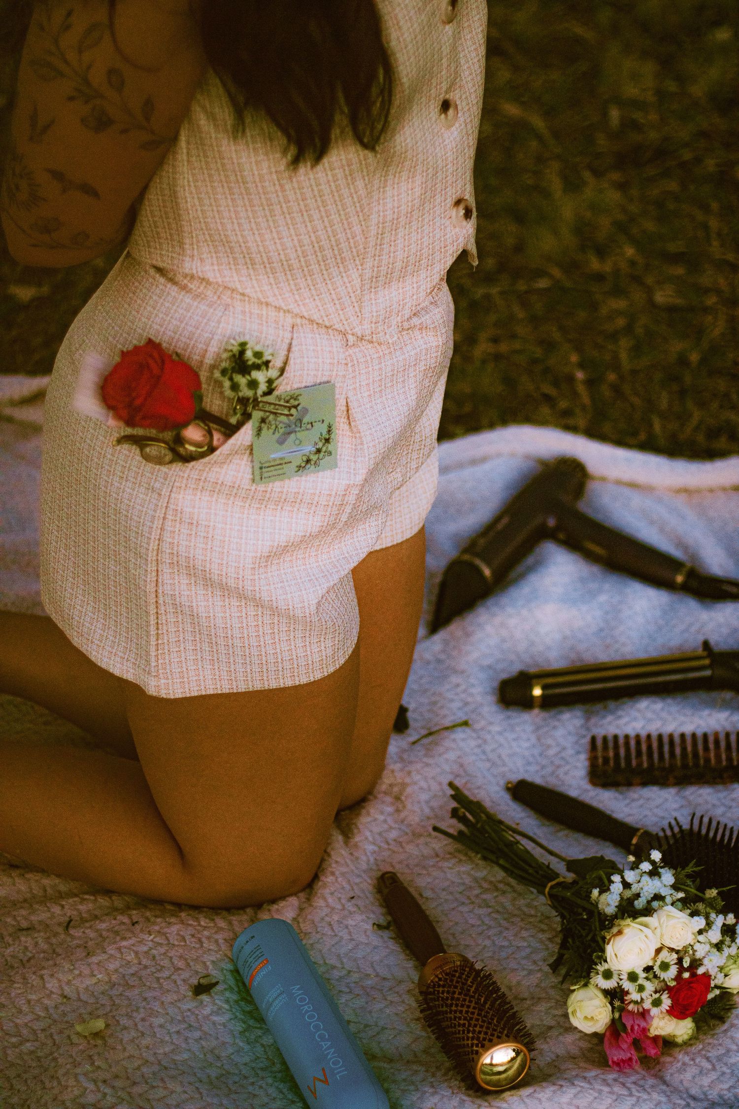 Woman in pink romper kneeling on blanket with hair tools, brushes, and floral accessories arranged beside her.