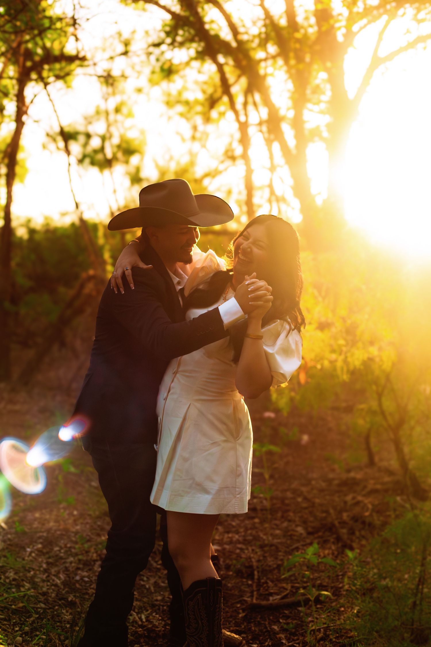 A couple embraces in golden sunset light in a forest, the man wearing a cowboy hat and the woman in a white dress.