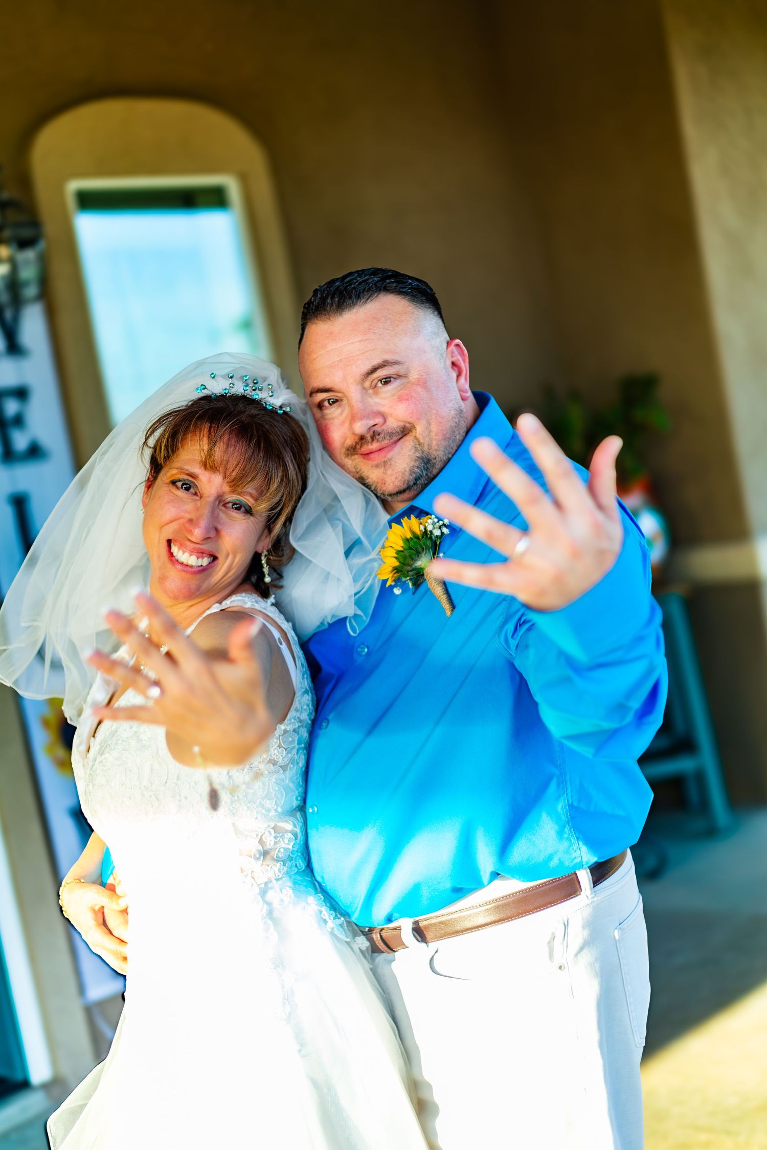 Smiling newlywed couple showing off wedding rings to camera, groom in blue shirt and bride in white lace dress.