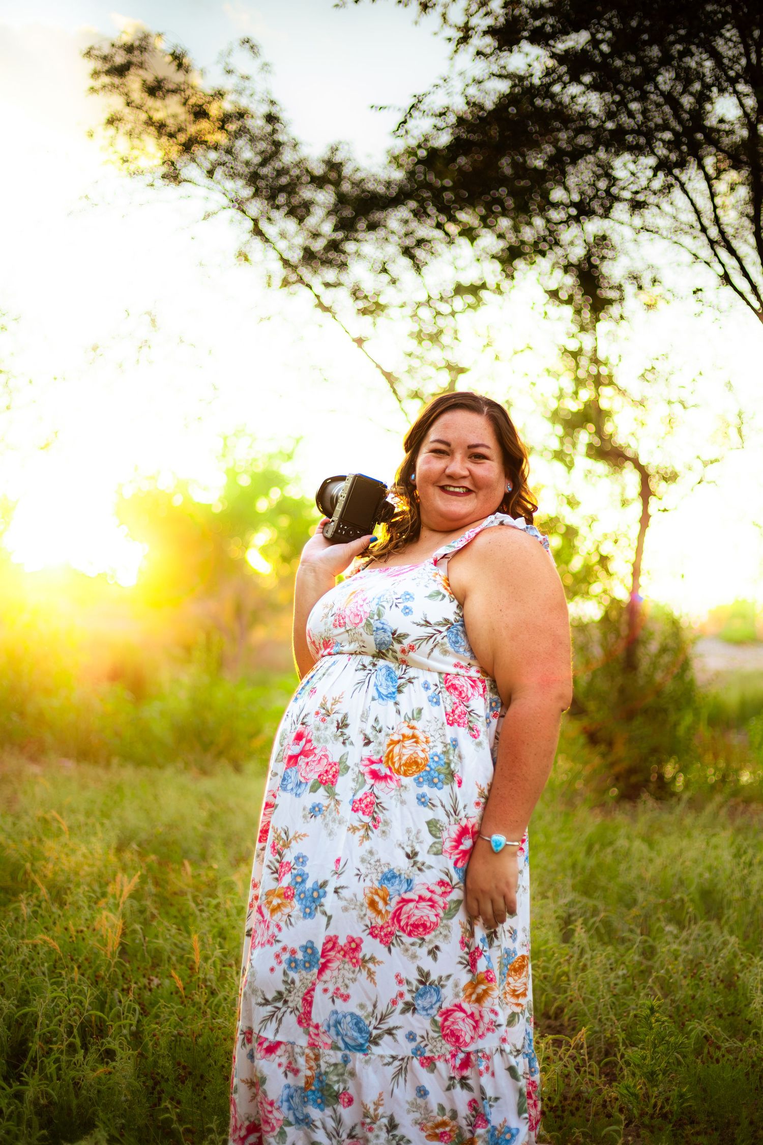 Pregnant woman in floral dress holding puppy outdoors at golden hour sunset.