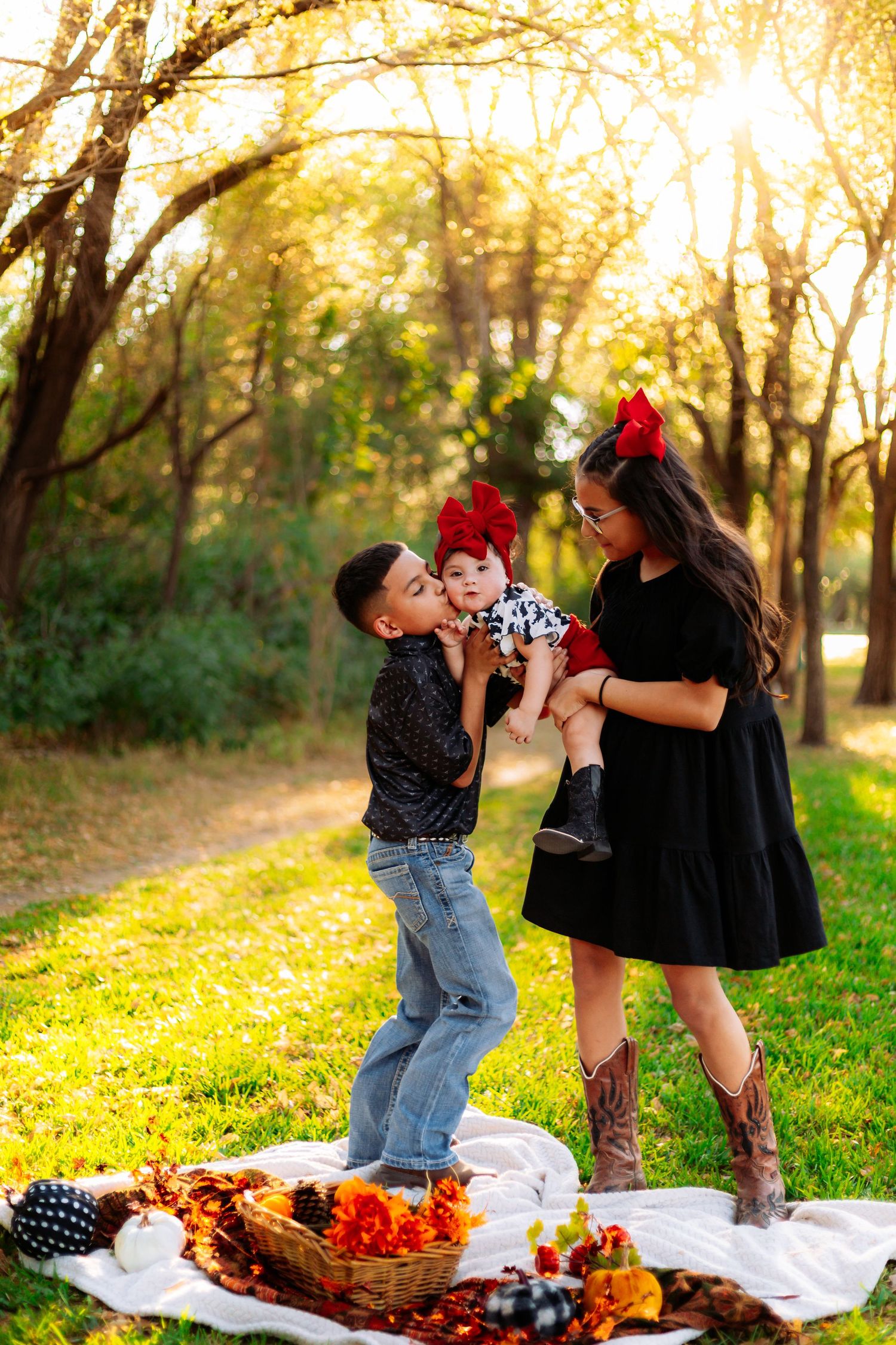 Family of three enjoying a fall picnic in a sunlit park, with older sibling kissing baby held by mom in black dress.