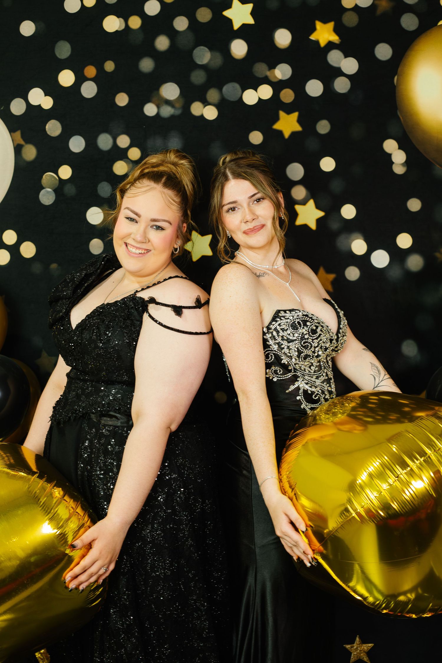 Two young women in elegant black prom dresses pose with gold balloons against a sparkling star backdrop.