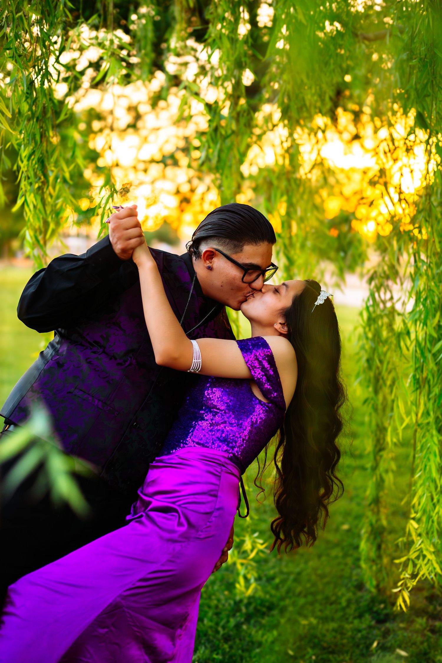 Couple sharing a romantic dip kiss under weeping willow tree, woman in purple gown and man in black suit.