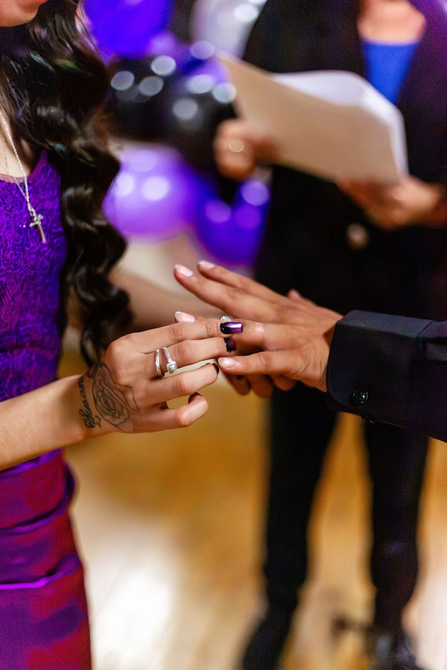 Close-up of a woman placing a wedding ring on a man's finger during a ceremony with purple balloons in the background.