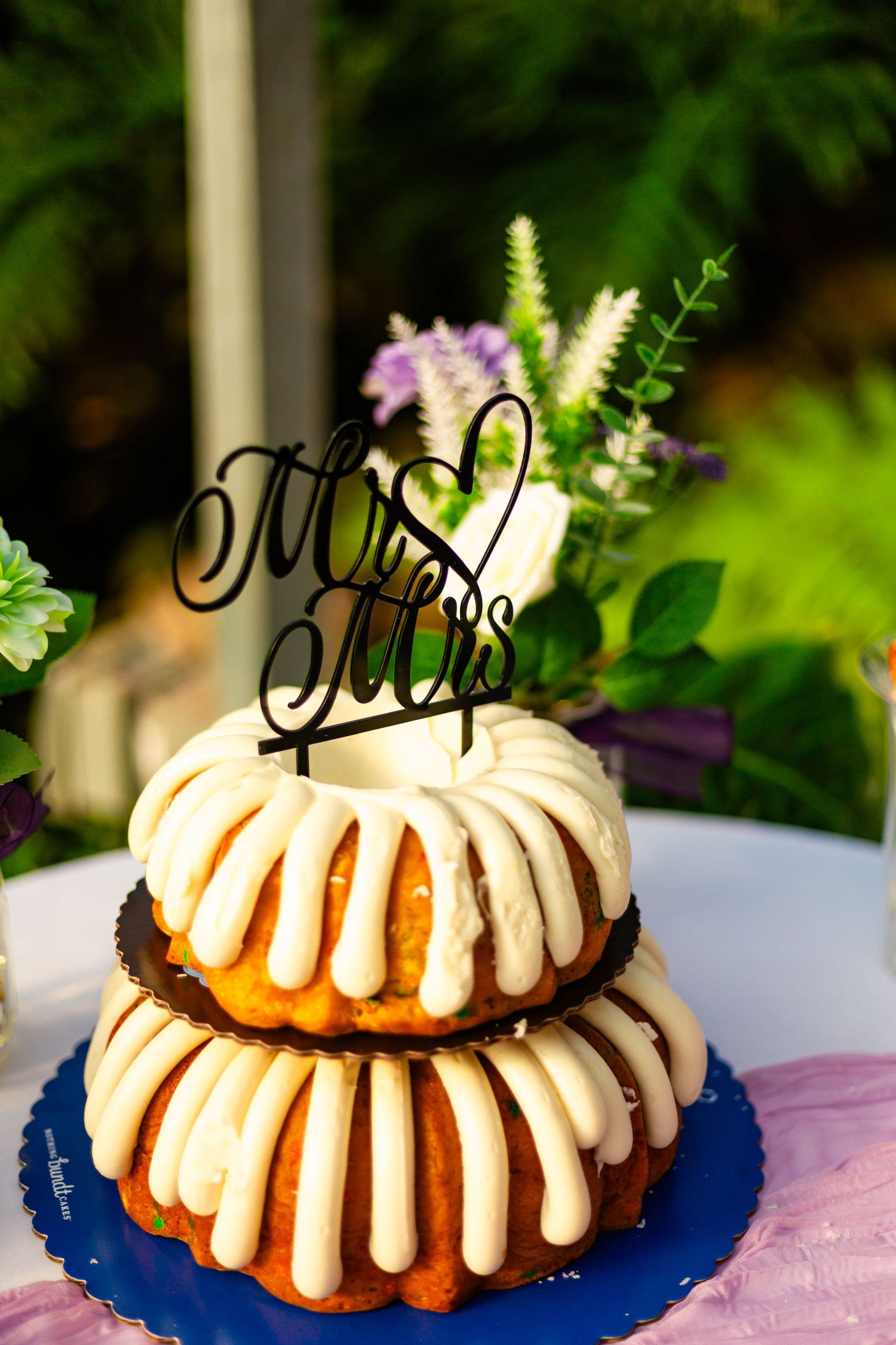 Two-tier bundt wedding cake with cream cheese frosting and Mr. and Mrs. cake topper on blue stand outdoors.