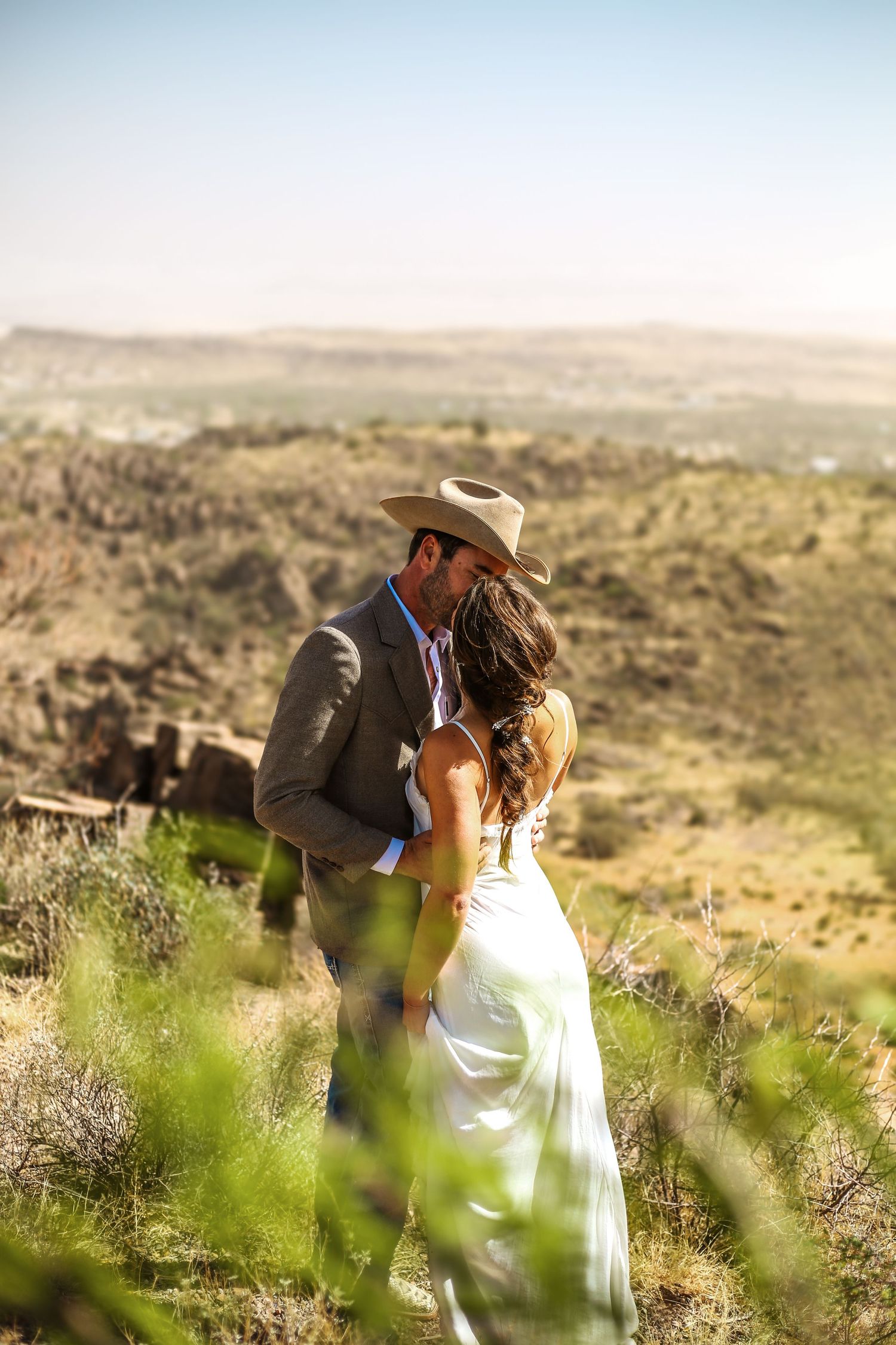 Couple sharing a romantic kiss in a desert landscape, groom in cowboy hat and suit, bride in white gown.