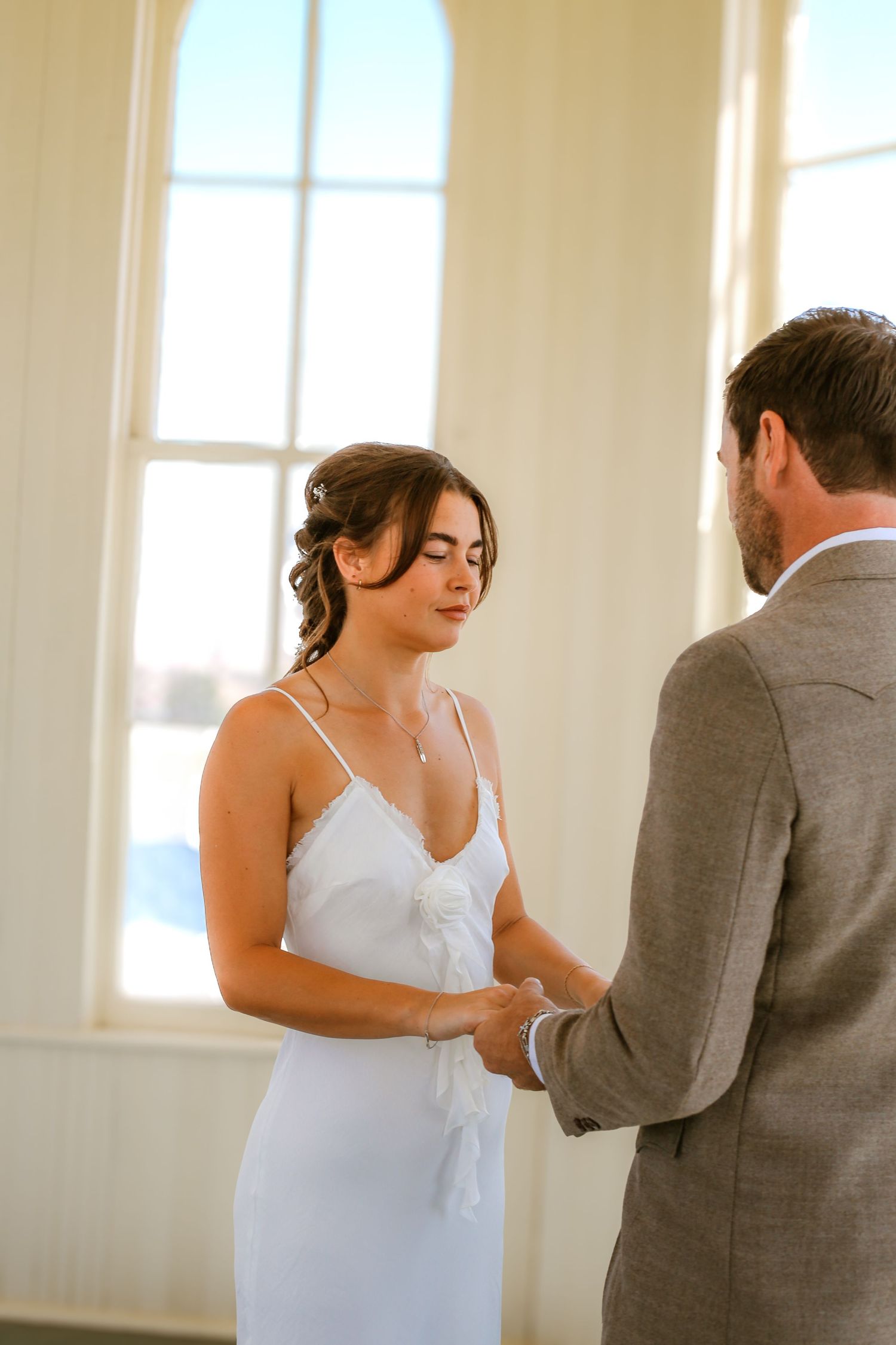 Bride in white slip dress holds groom's hands during intimate indoor wedding ceremony near sunlit window.