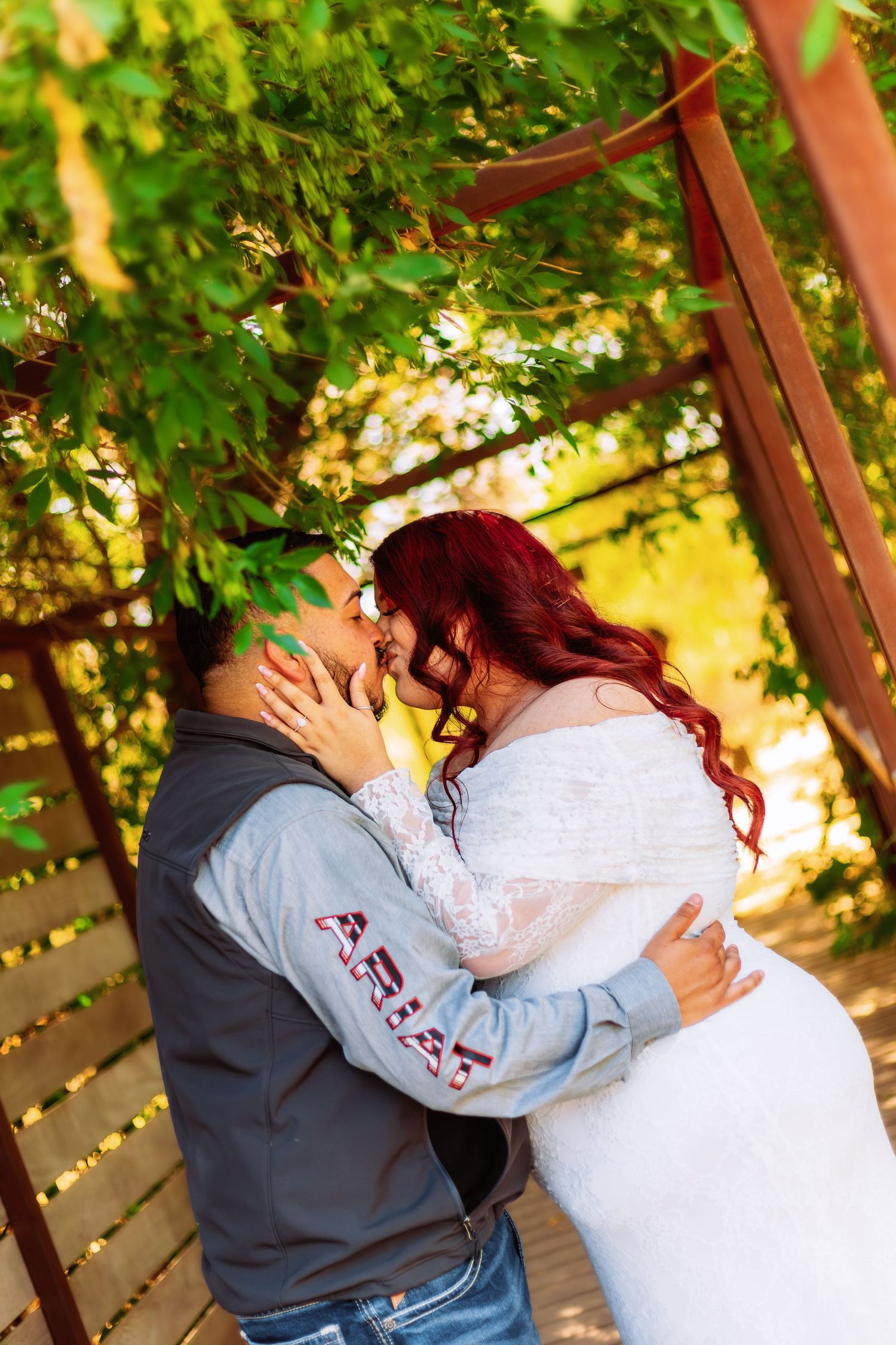 A bride in a white lace gown and groom share a romantic kiss under a wooden pergola with lush green foliage.