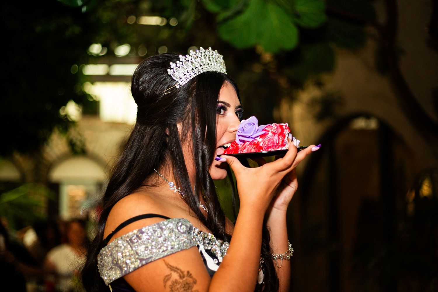 Young woman wearing a sparkling tiara and off-shoulder gown kisses a small gift box at a quinceañera celebration.