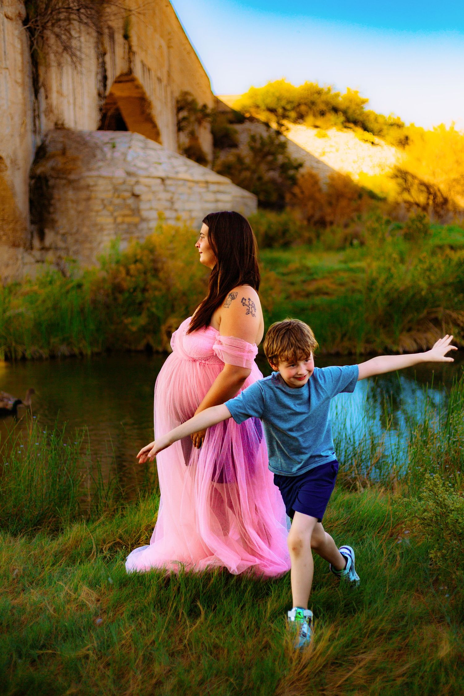 Pregnant woman in pink gown with young boy outdoors near rocky cliffs and pond at golden hour.