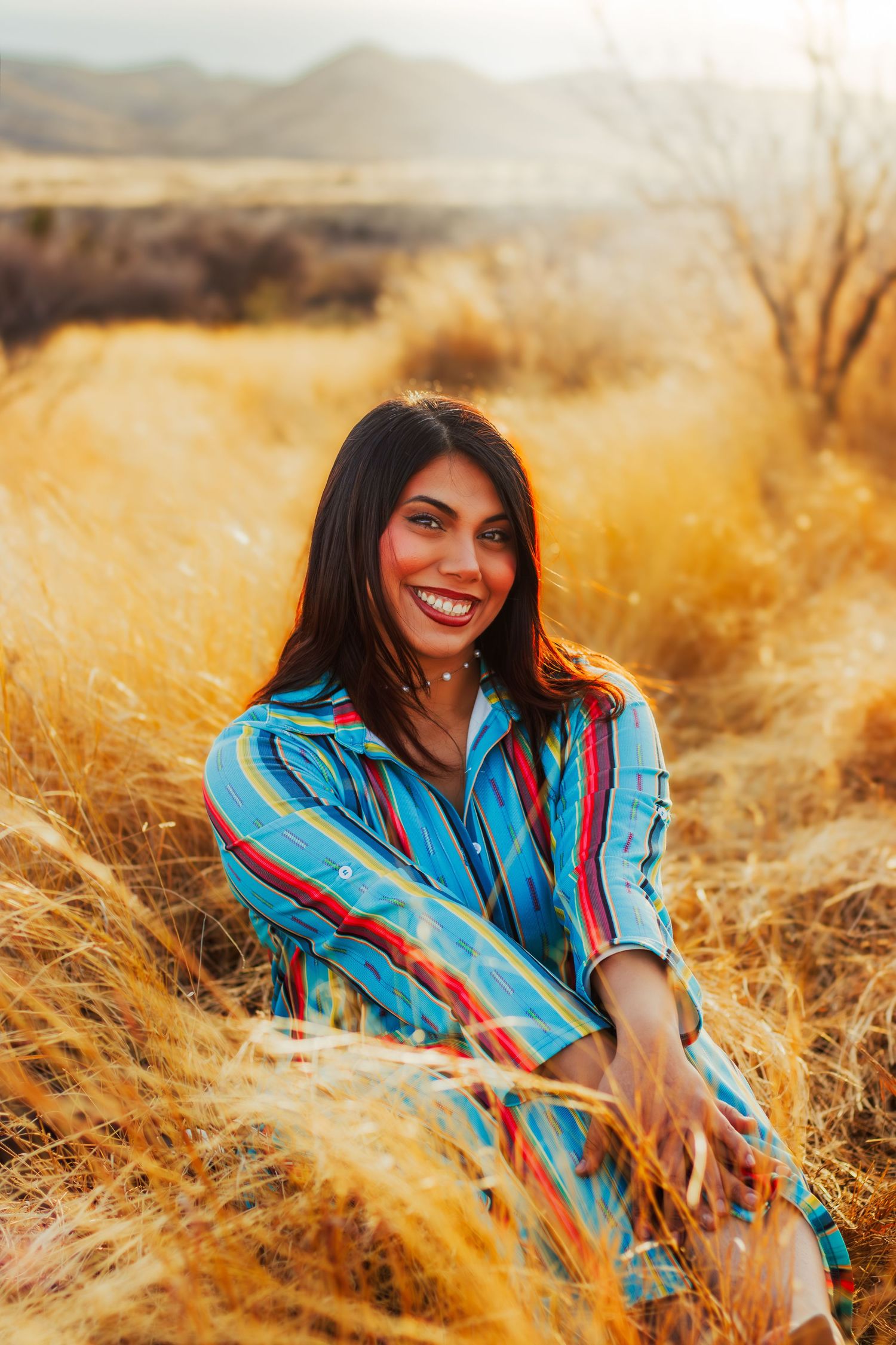 Smiling woman in colorful striped outfit sitting in golden dry grass field at sunset.