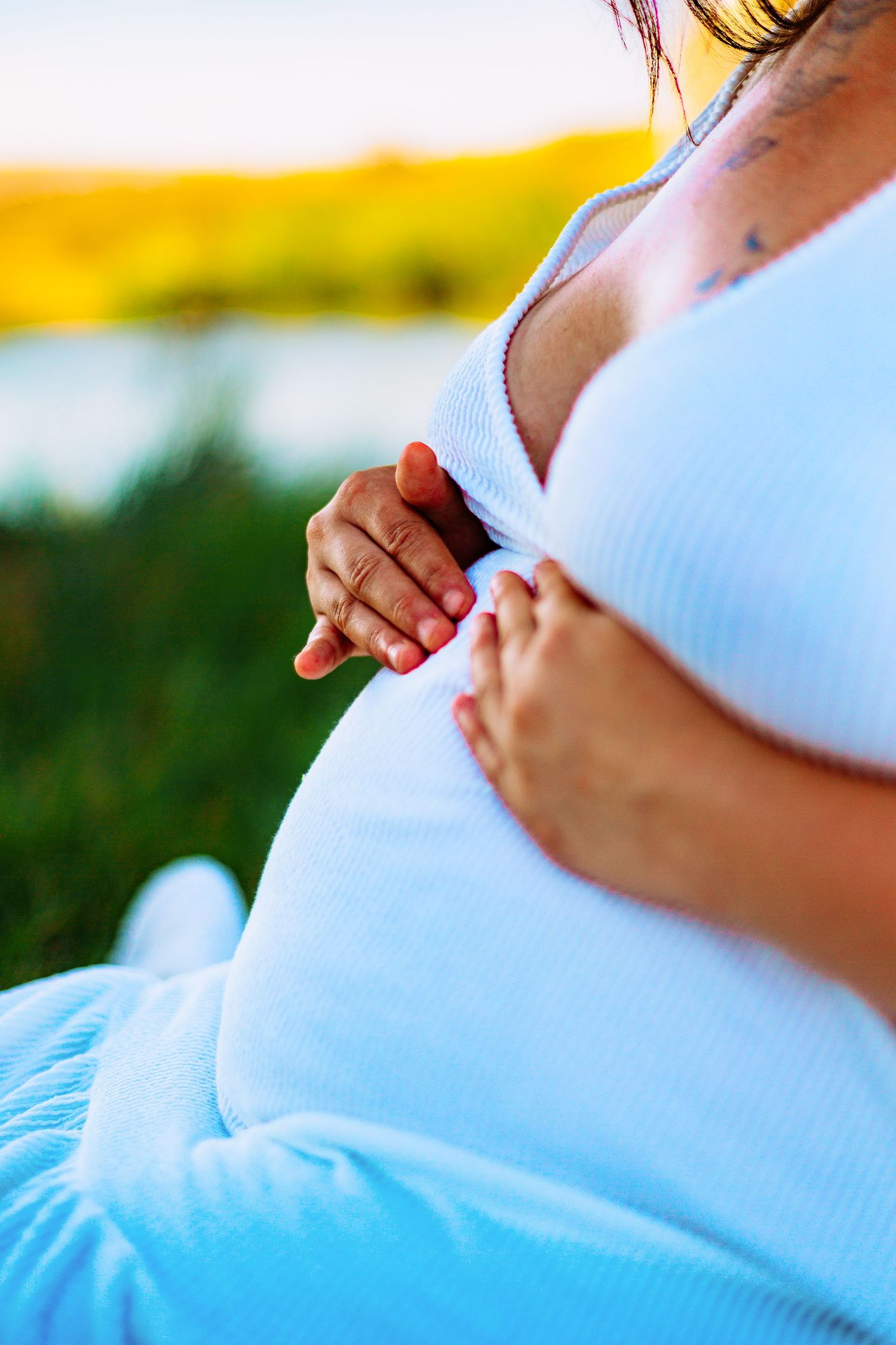 Pregnant woman in white top and blue pants cradling her baby bump outdoors near a lake at golden hour.