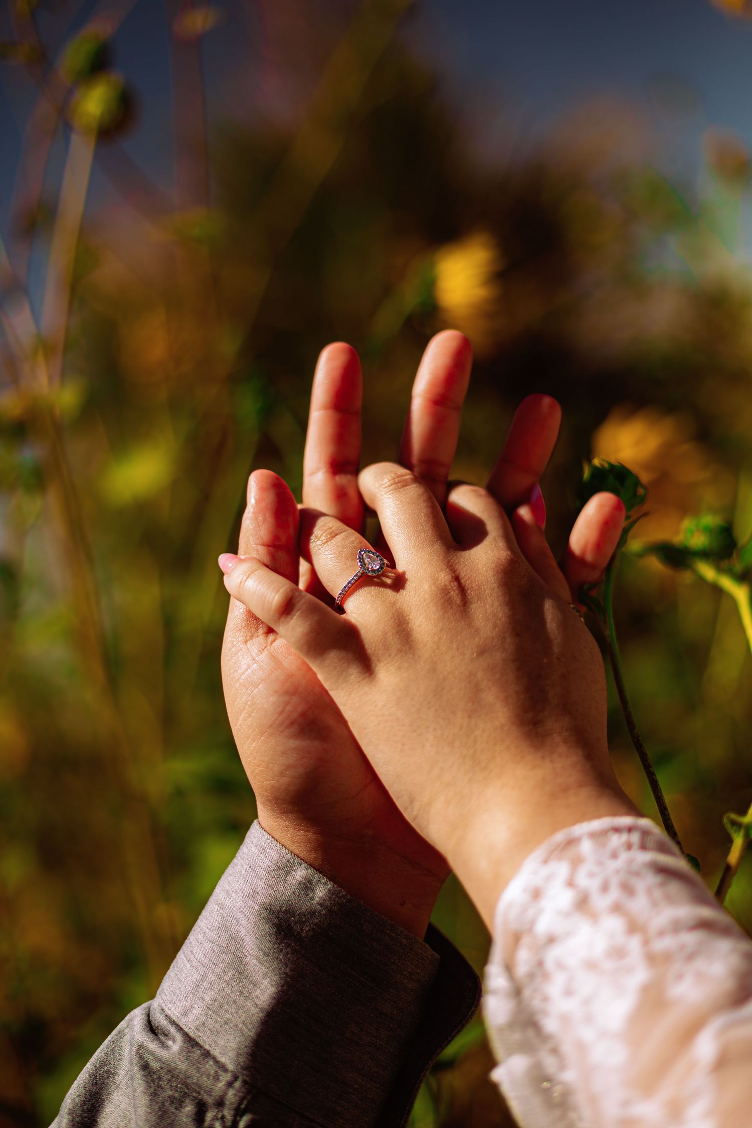 Two hands touching with an engagement ring on finger, set against a blurred natural outdoor background.