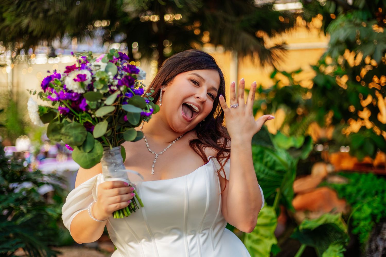 Joyful bride in white dress shows off ring, holding purple bouquet, laughing outdoors among lush greenery.