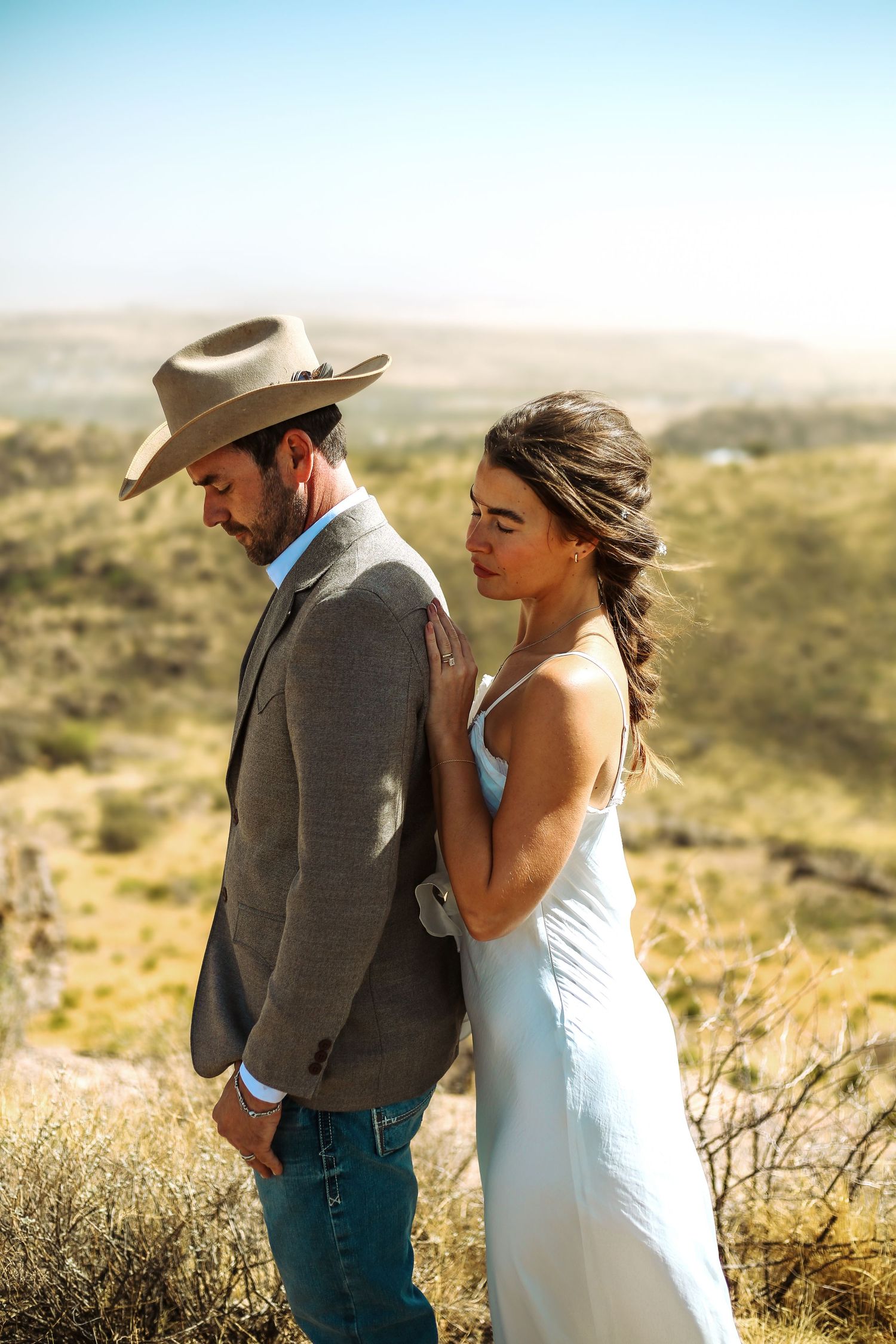 A bride in a white dress leans against a groom in a cowboy hat and gray suit in a scenic desert landscape.